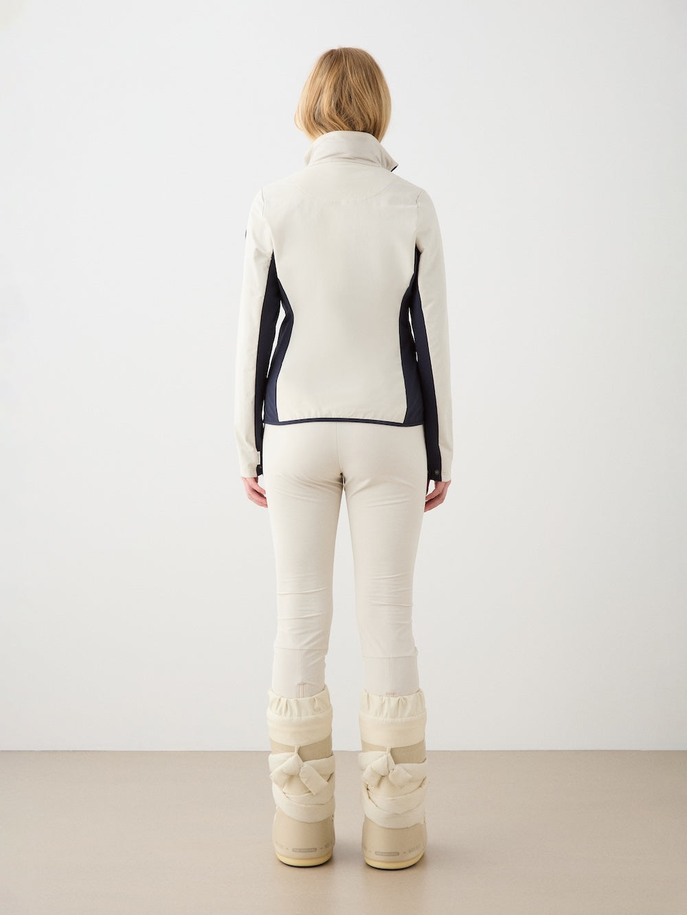 A person with short blonde hair faces away, wearing the We Norwegians Beito Jacket Women in cream with matching pants featuring black side panels and large cream winter boots, set against a plain light background.