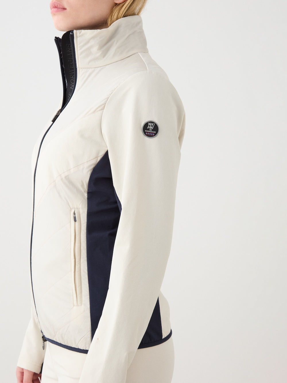 A woman stands against a white background wearing the We Norwegians Beito Jacket Women in cream, featuring black side panels, a black zipper, high collar, and a circular logo patch on the sleeve.