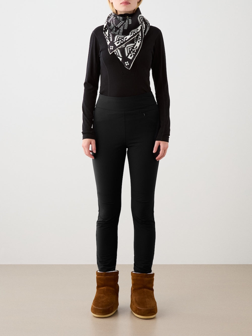 A person faces forward on a neutral background, wearing a black long-sleeve top, We Norwegians Beito Pant Women outdoor training pants, a black-and-white patterned scarf, and brown suede ankle boots.