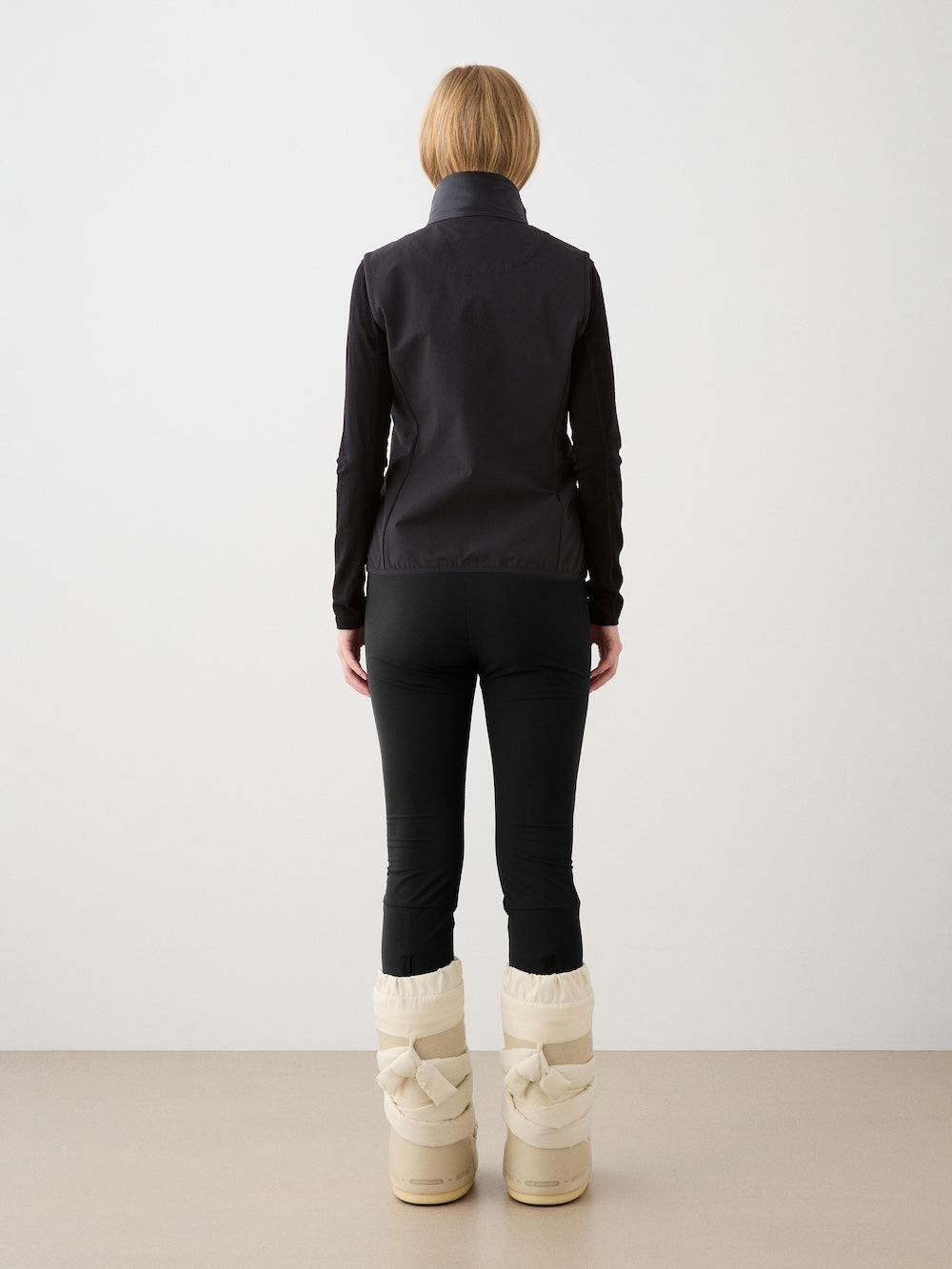 A person with short blonde hair, seen from behind, wears a black jacket, black pants, large cream puffy boots, and the We Norwegians Beito Vest Women, standing against a plain light background.