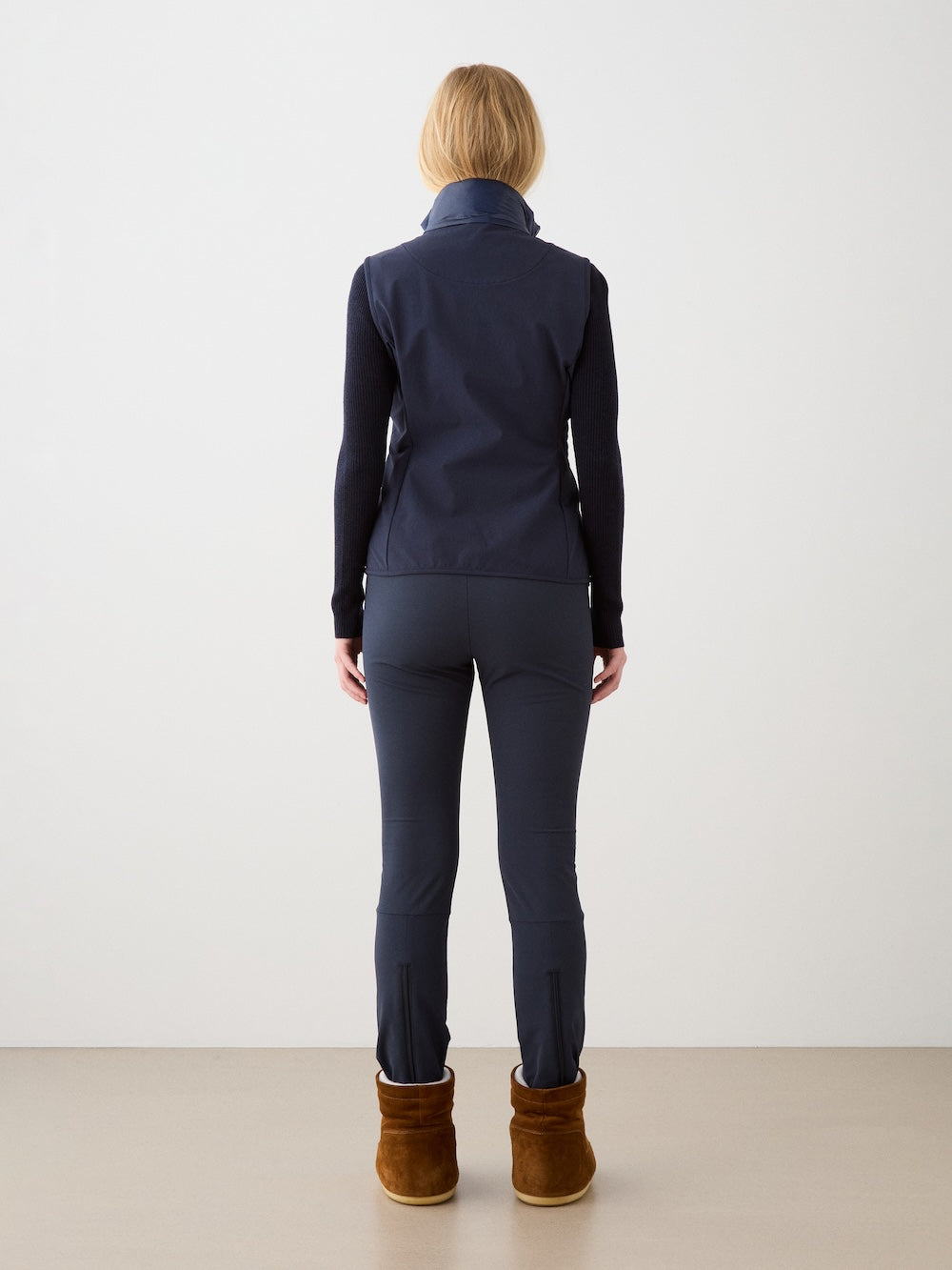 A woman with shoulder-length blonde hair, seen from behind, wears a We Norwegians Beito Vest layered under a navy jacket, fitted pants, and brown ankle boots against a light background.