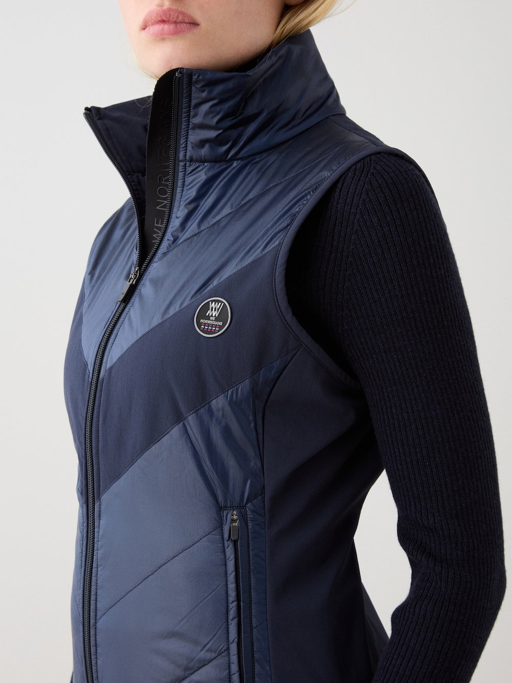 A woman wears the We Norwegians Beito Vest Women in navy, featuring a circular logo on the chest and a non-fluorinated DWR finish, layered over a dark ribbed sweater, posed against a plain light background.