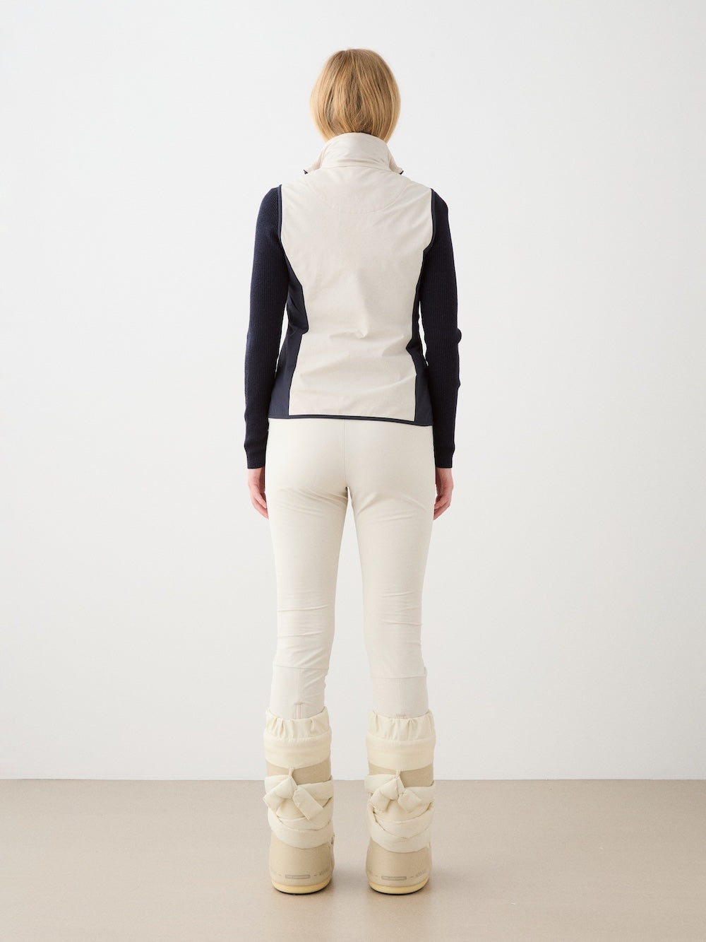 A woman with light brown hair, seen from behind, wears the We Norwegians Beito Vest Women over a dark long-sleeve top, cream pants, and beige winter boots, standing against a plain light background.