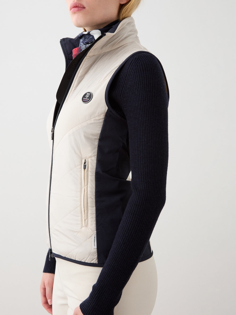 A woman models the We Norwegians Beito Vest Women—a cream and black merino wool zip-up with non-fluorinated DWR—over a black long-sleeve top and cream pants, standing in profile against a plain light background.