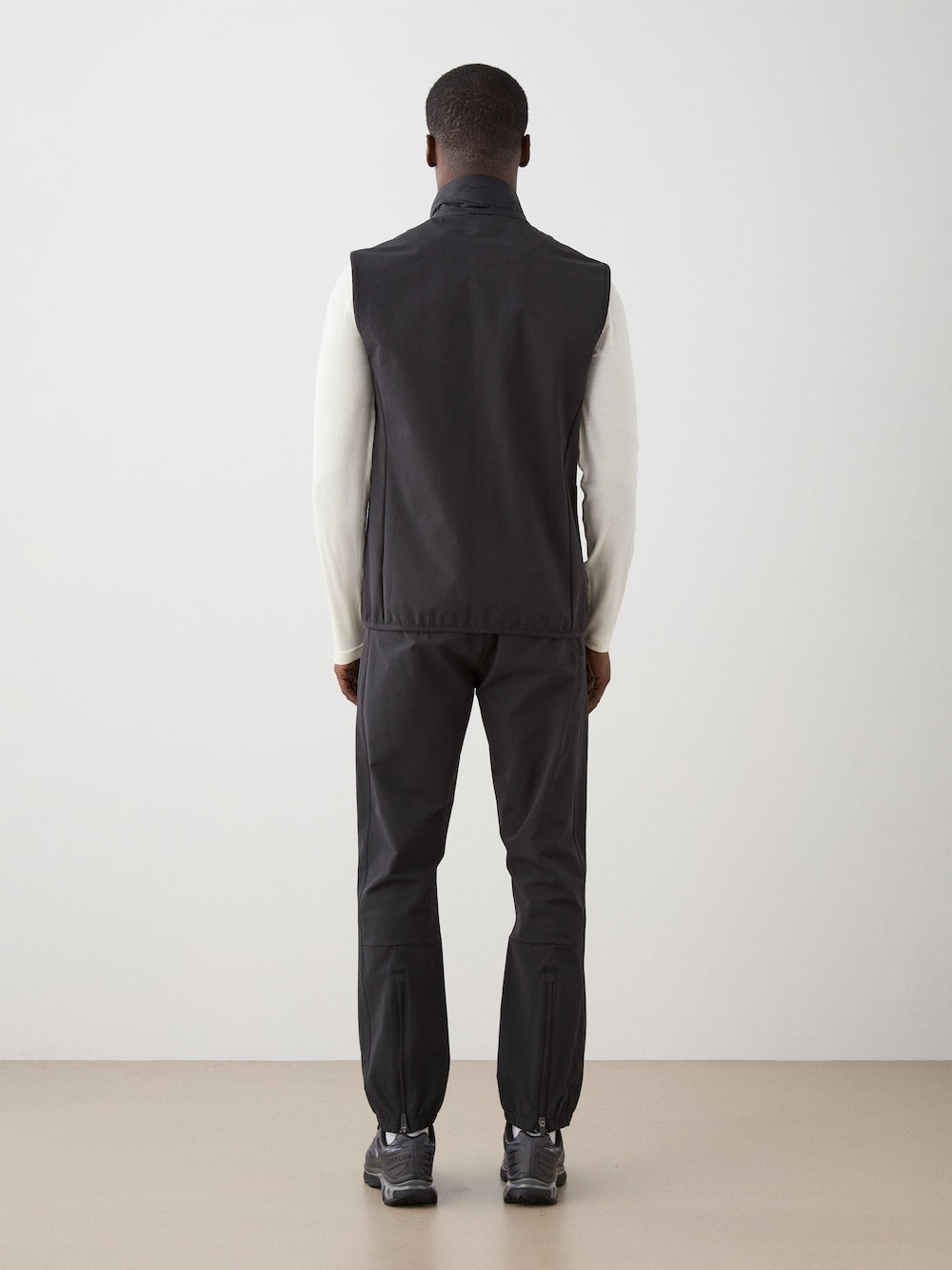 A person faces away from the camera, wearing the We Norwegians Beito Vest Men in black over a white long-sleeve shirt, with black pants and sneakers, set against a plain minimal indoor background—ideal for performance layering.