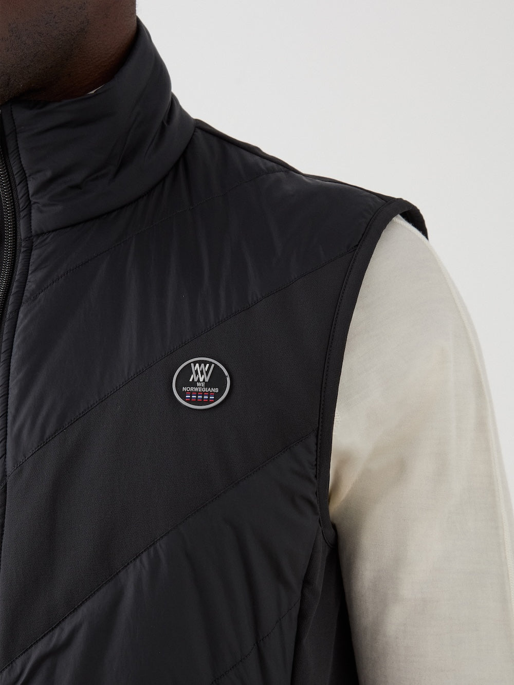 A close-up shows a person wearing the black Beito Vest Men by We Norwegians over a light long-sleeve shirt. The quilted vest features a small circular logo patch on the chest, with the person's face out of frame.