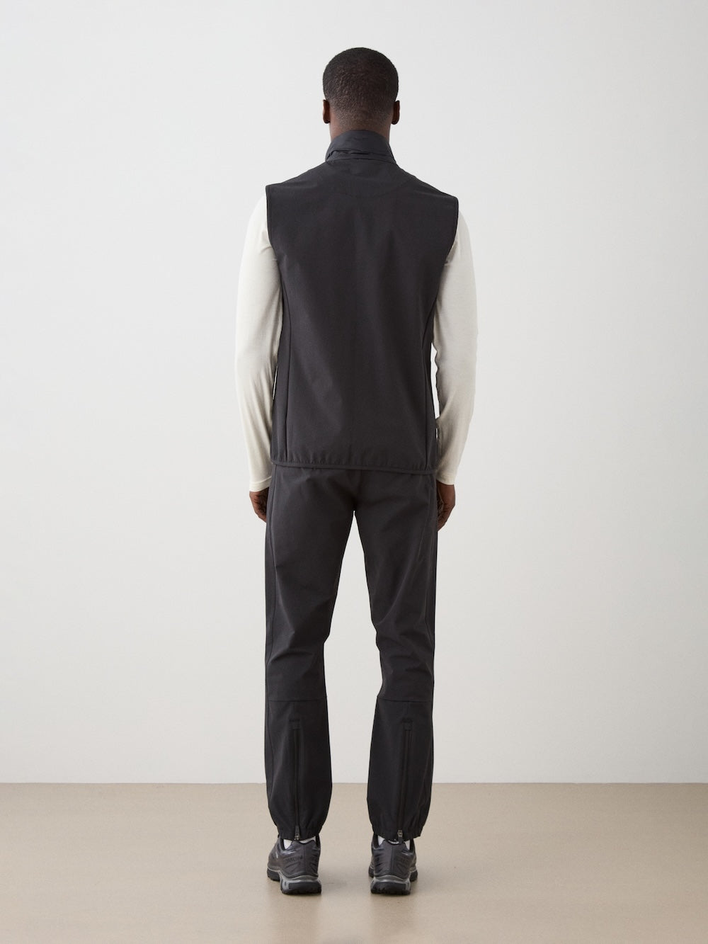 A man faces away from the camera, wearing a We Norwegians Beito Vest Men in black over a white long-sleeve shirt, with black pants and sneakers—ideal performance layering against a plain light background.