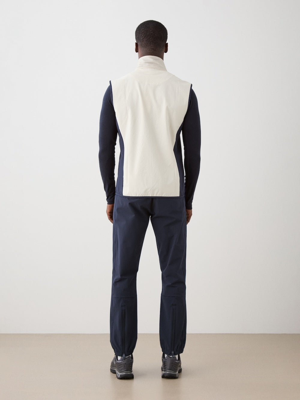 A man faces away, wearing the We Norwegians Beito Vest Men—a white merino wool sleeveless vest—layered over a dark long-sleeve shirt, with dark blue pants featuring elastic cuffs and dark sneakers, in a minimalist indoor setting.