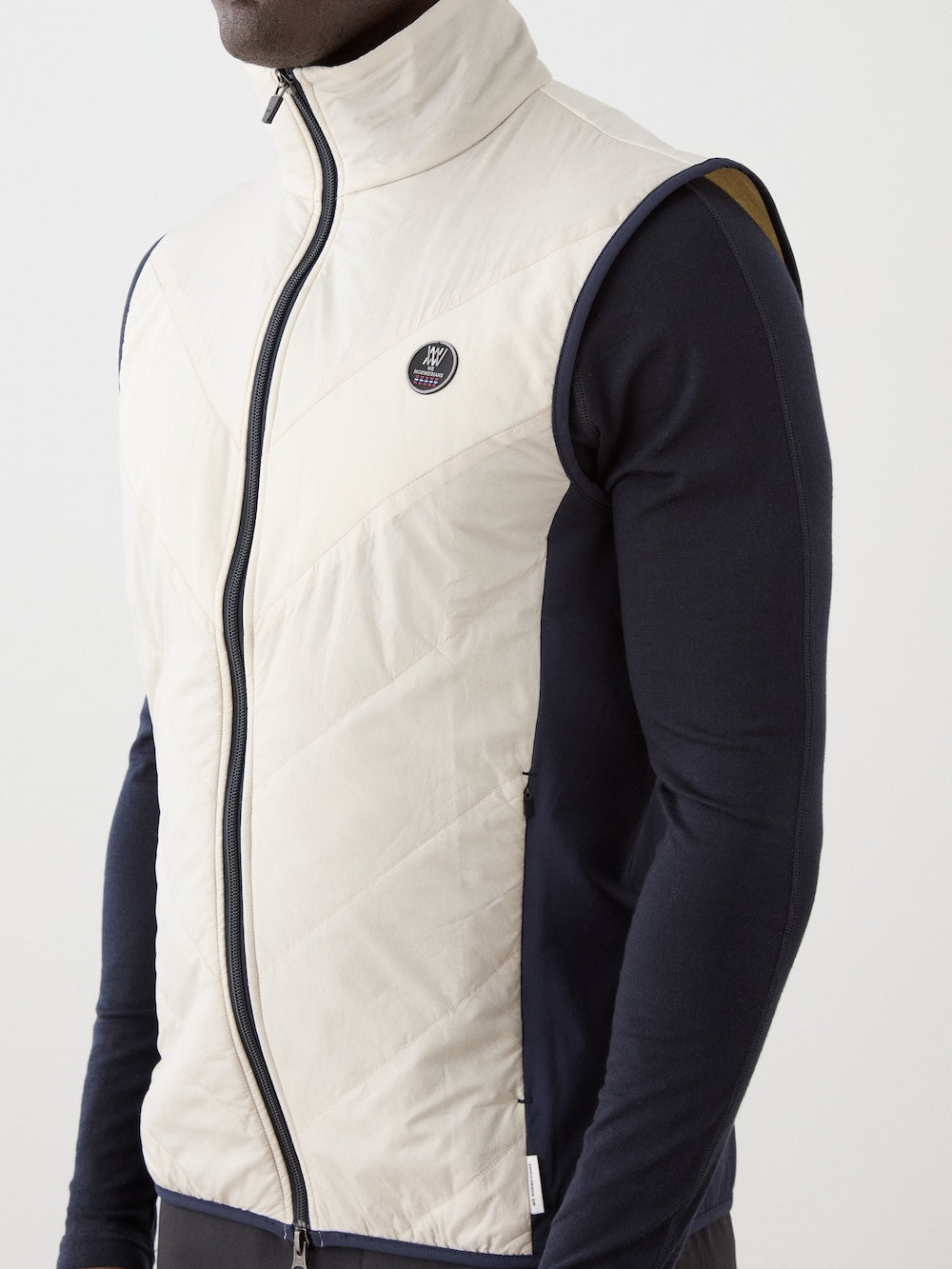 A person wears the cream Beito Vest Men by We Norwegians—a sleeveless merino wool layer with a round black and white patch logo—over a fitted black long-sleeve shirt, ideal for performance layering. Shown from shoulders to waist.