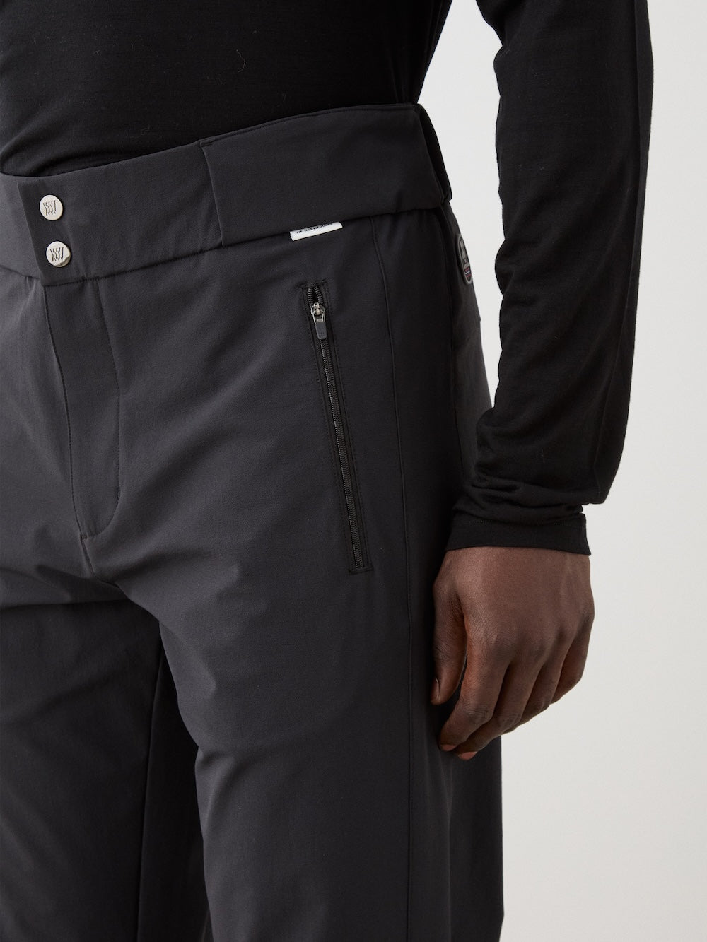 A person wears We Norwegians Beito Pant Men in black with zippered side pockets and two visible waistband buttons, paired with a black long-sleeve shirt, right hand resting by their side.
