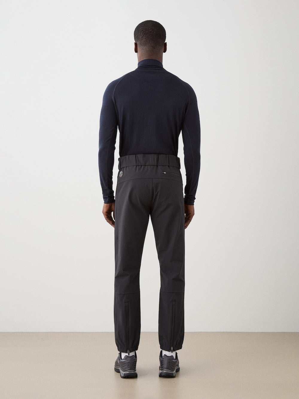 A man faces away from the camera in a minimal studio, wearing a fitted black long-sleeve top, We Norwegians Beito Pant Men made of dark merino wool with elastic ankles, and black sneakers. The background is plain and light.