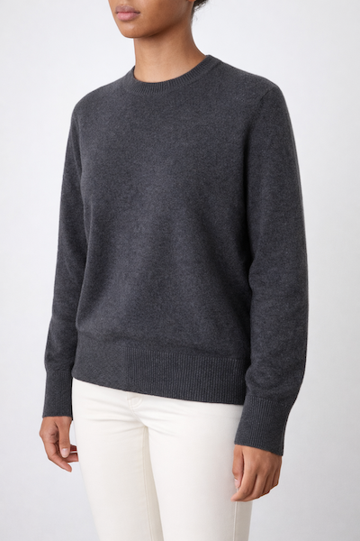 A person wears the We Norwegians Blefjell Crewneck Women—an oversized dark gray 100% cashmere sweater with ribbed cuffs and hem—paired with white pants, standing against a plain light background. The face is not visible.