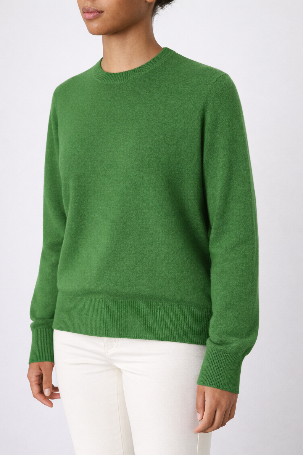 A person wears the We Norwegians Blefjell Crewneck Women - Pure Cashmere sweater in green with off-white pants, standing against a light gray background. Their head is partly out of the frame.