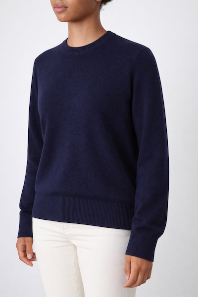 A person is shown from neck to knees, wearing the We Norwegians Blefjell Crewneck Women - Pure Cashmere sweater in dark navy blue with long sleeves, paired with off-white pants, standing against a plain light background.