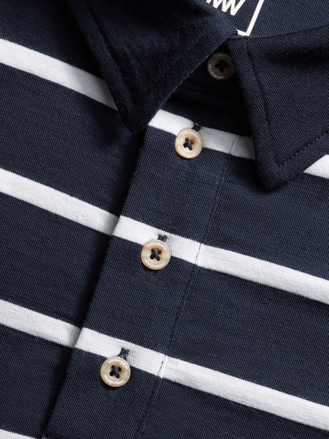 Close-up of the Kragero Polo Men Navy by We Norwegians. This navy blue polo shirt features white horizontal stripes, a dark collar, and three beige buttons down the center. Made from Merino wool, this comfortable and textured fabric is perfect for summer activities.