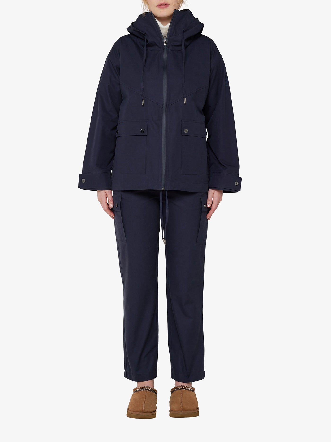A person stands wearing the We Norwegians Lyngen Jacket Women in Navy Blue, featuring a hooded design with multiple pockets and crafted from weatherproof fabric, paired with matching navy pants. They complete the outfit with brown slippers. The ensemble presents a casual and relaxed look reminiscent of RAF pilots against a plain white background.