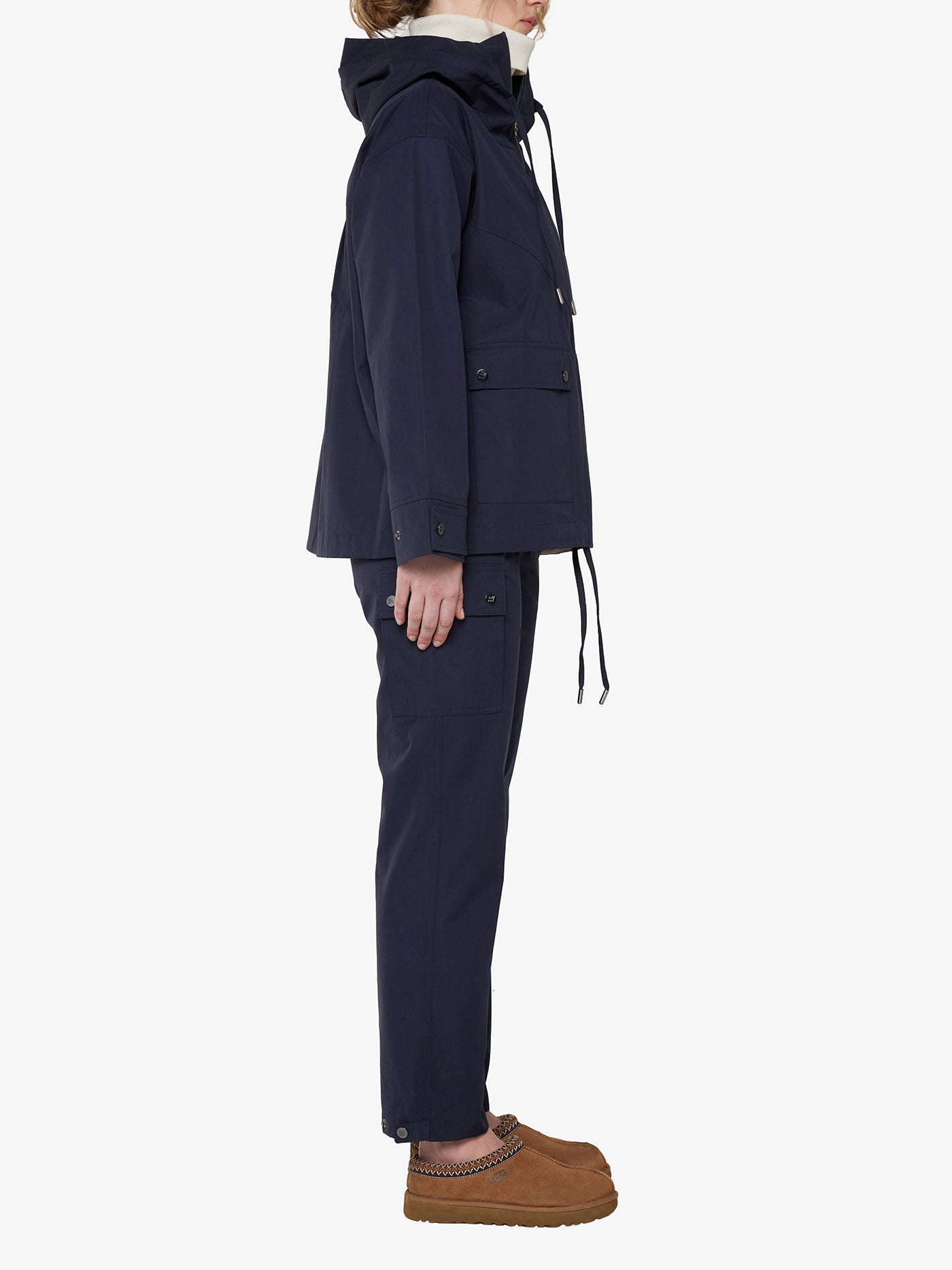 A person is standing sideways wearing the Lyngen Jacket Women Navy Blue by We Norwegians, made of Ventile® cotton, and matching pants with buttoned pockets. They have tan slip-on shoes with a thick sole. The weatherproof jacket features a hood inspired by those worn by RAF pilots, and the person is facing to the left against a plain white background.