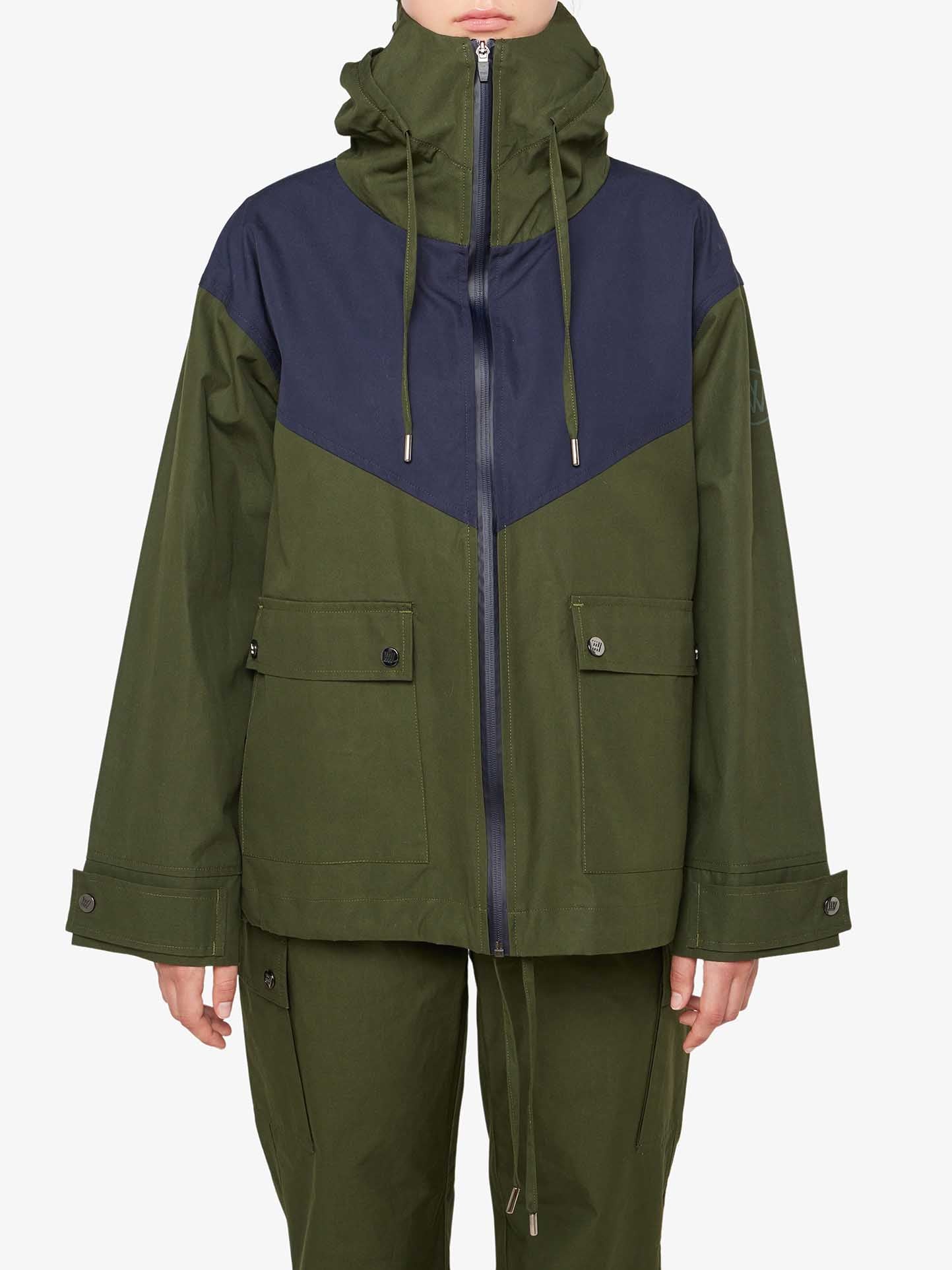 A person is wearing the We Norwegians Lyngen Jacket Women in Olive Green, a green and navy color-blocked jacket with a hood and zip-up front, crafted from Ventile® cotton fabric. The jacket features two large pockets with buttons and drawstring cords hanging from the hood. The person, an outdoor enthusiast, is also wearing green pants. The background is plain white.