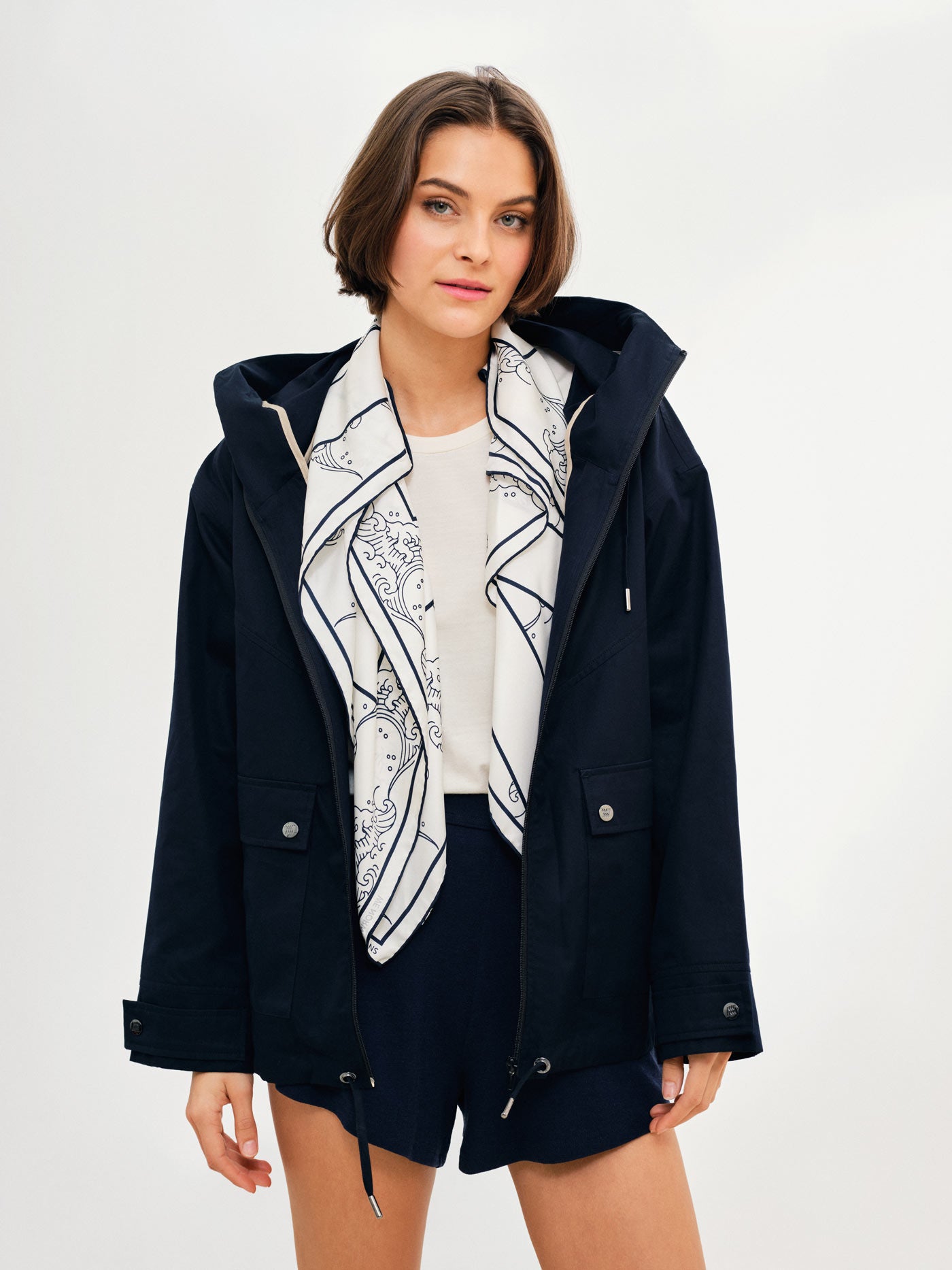 A woman with short brown hair sports the Lyngen Jacket for Women in Navy Blue by We Norwegians, layered over a white shirt and paired with matching navy blue shorts. She accessorizes with a white and blue patterned scarf around her neck. The plain white background provides a clean and simple setting.