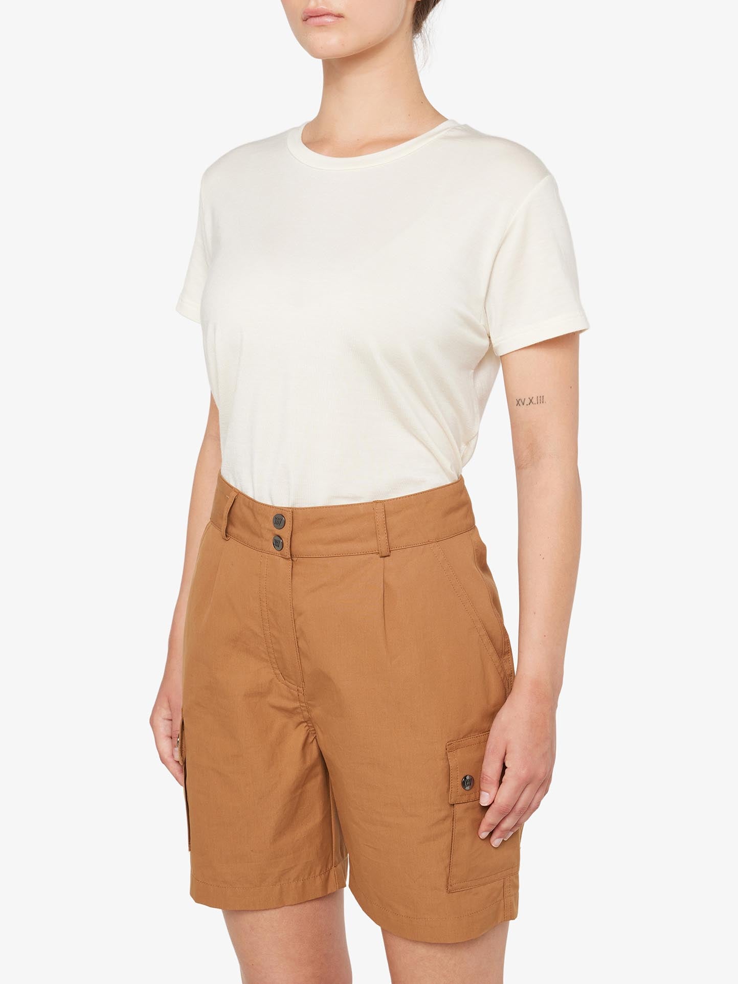 A person is wearing a plain white t-shirt tucked into high-waisted, knee-length Lyngen Shorts Women Cinnamon from We Norwegians, featuring side pockets and button closures that make them ideal for outdoor enthusiasts. The background is white, and a small tattoo is visible on the person's left forearm.