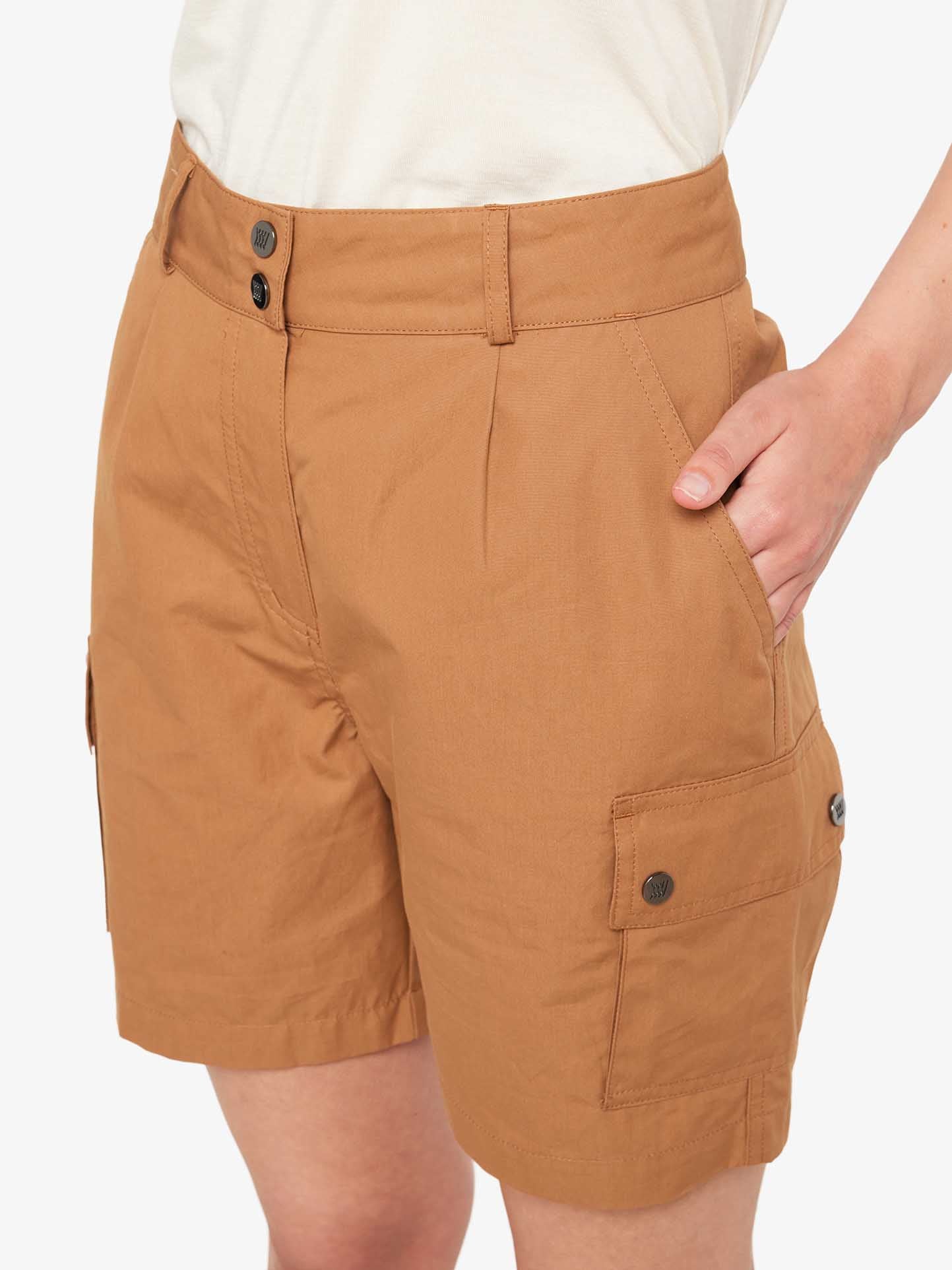 A person is wearing We Norwegians' Lyngen Shorts Women in Cinnamon, designed with side pockets and button closures, making them perfect for outdoor enthusiasts. Their left hand rests in one of the front pockets. Made from sturdy, weatherproof fabric, these shorts are paired with a white top, showing only part of the person's torso and legs.