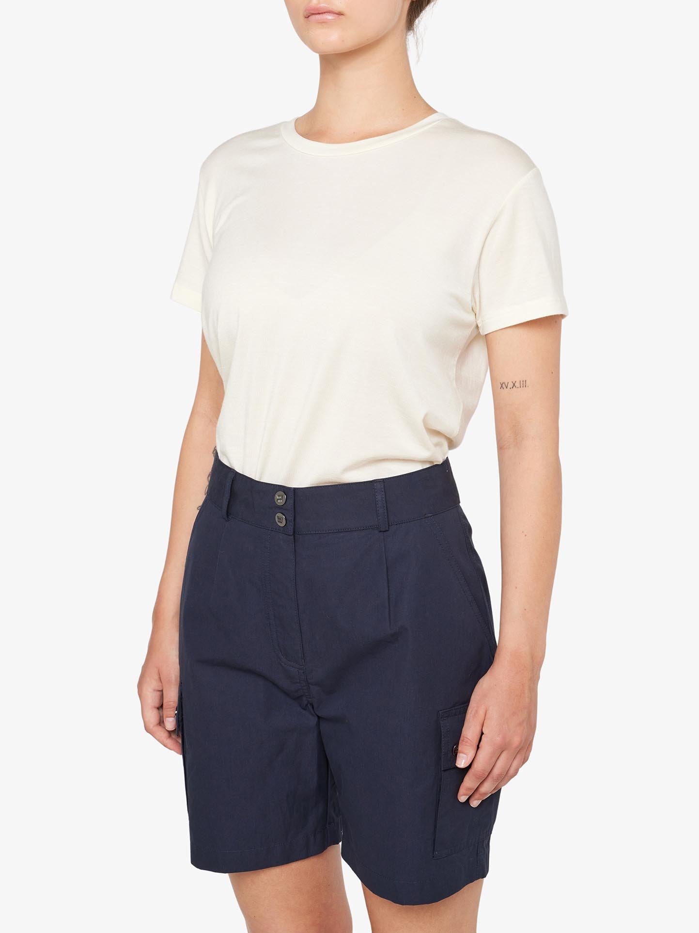 A person stands against a plain white background wearing a cream-colored short-sleeve t-shirt tucked into We Norwegians' Lyngen Shorts Women Navy Blue. The weatherproof shorts, perfect for outdoor enthusiasts, feature pockets and button details. The person has a tattoo with Roman numerals on the upper part of their left arm.