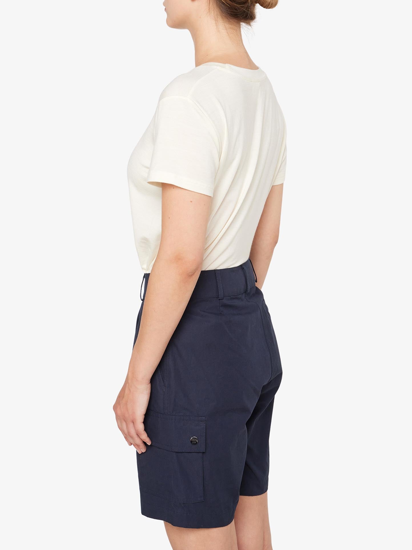 A person with light skin is standing sideways, facing left, wearing a white t-shirt tucked into We Norwegians' Lyngen Shorts for women in navy blue. The shorts are crafted from Ventile® fabric and feature side pockets. The plain white background highlights the outfit's weatherproof qualities, ideal for outdoor enthusiasts.