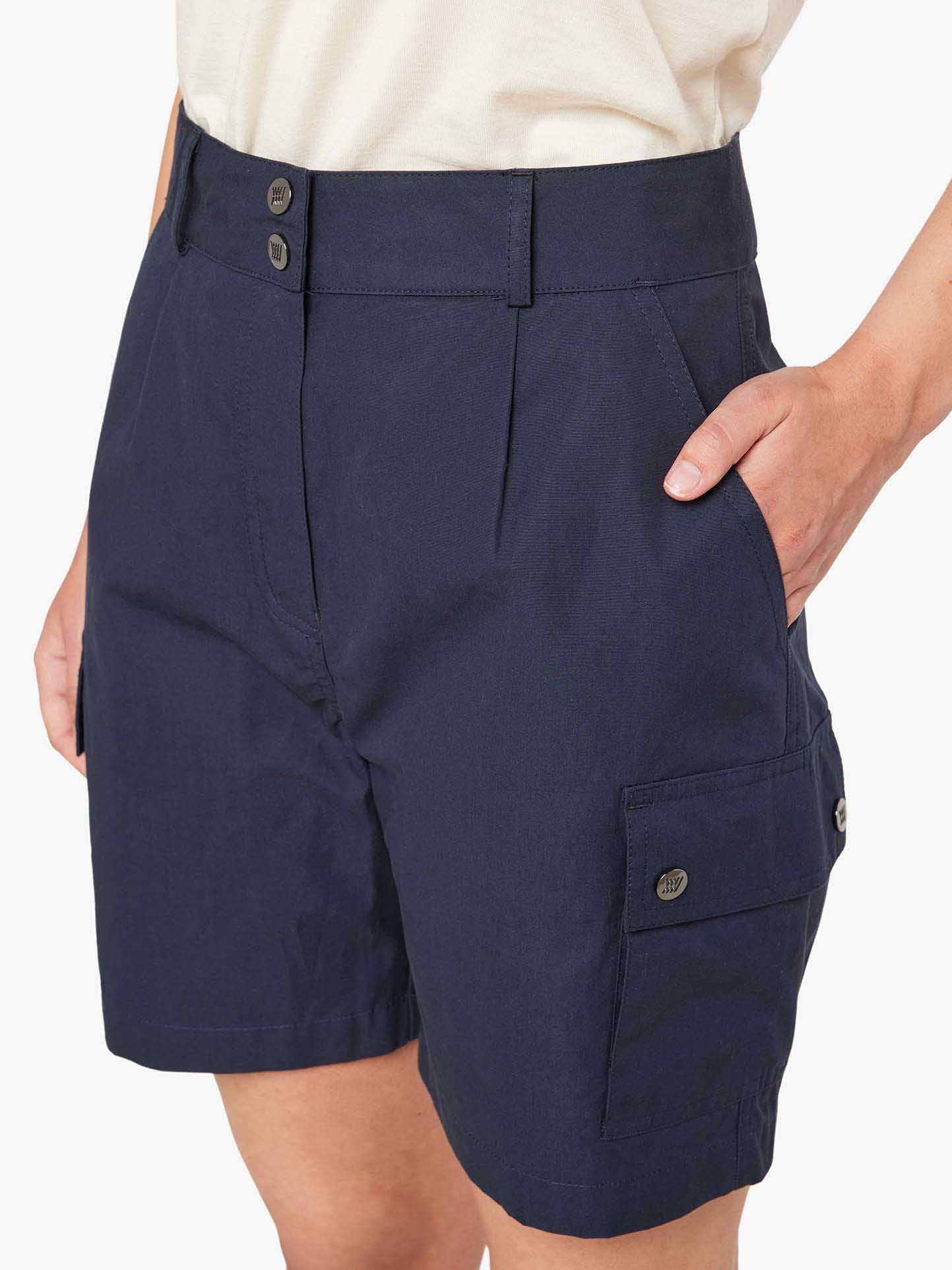 A woman wearing the We Norwegians Lyngen Shorts Women in Navy Blue, with her hands in the pockets. Made from Ventile® fabric for superior weatherproof qualities, these shorts feature two front buttons and a pocket flap button on the side. Paired with a white tucked-in shirt, this outfit is perfect for outdoor enthusiasts. The background is plain white.