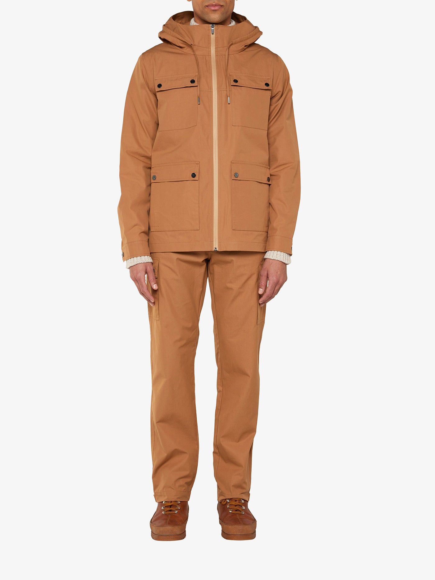 A person stands against a white background wearing the We Norwegians Lyngen Jacket Men Cinnamon, featuring a hood and four front pockets, paired with matching brown pants. Made from weatherproof Ventile® cotton fabric, this outfit is ideal for cool weather. They are also wearing brown shoes, completing the casual, functional look.
