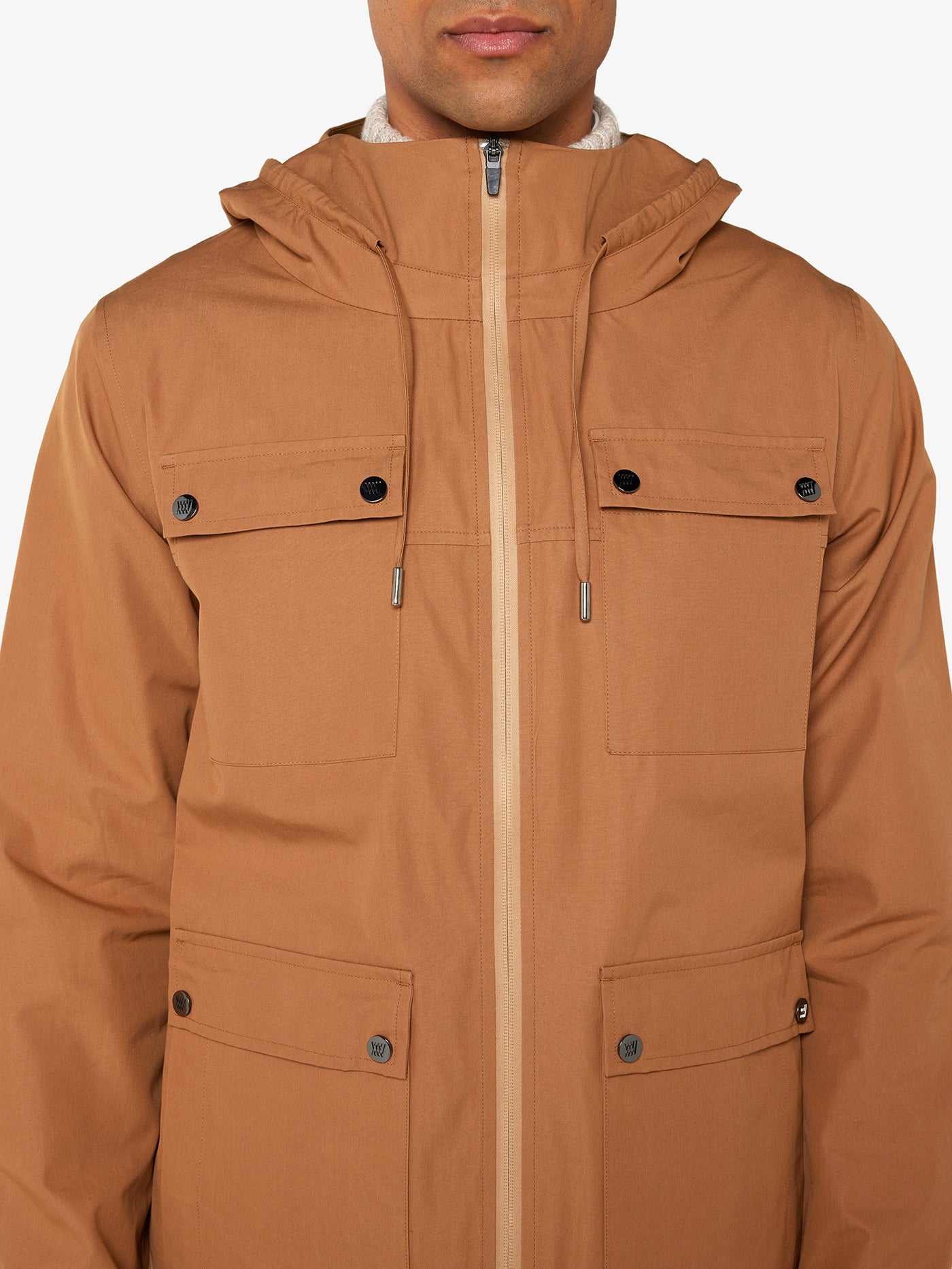 A person is wearing the Lyngen Jacket in Cinnamon by We Norwegians, made from Ventile® cotton fabric with buttoned pockets on the chest and near the waist. The weatherproof qualities are evident, featuring a front zipper closure. Only the lower half of their face and upper torso are visible, highlighting the detailed features of this RAF pilots-inspired jacket.