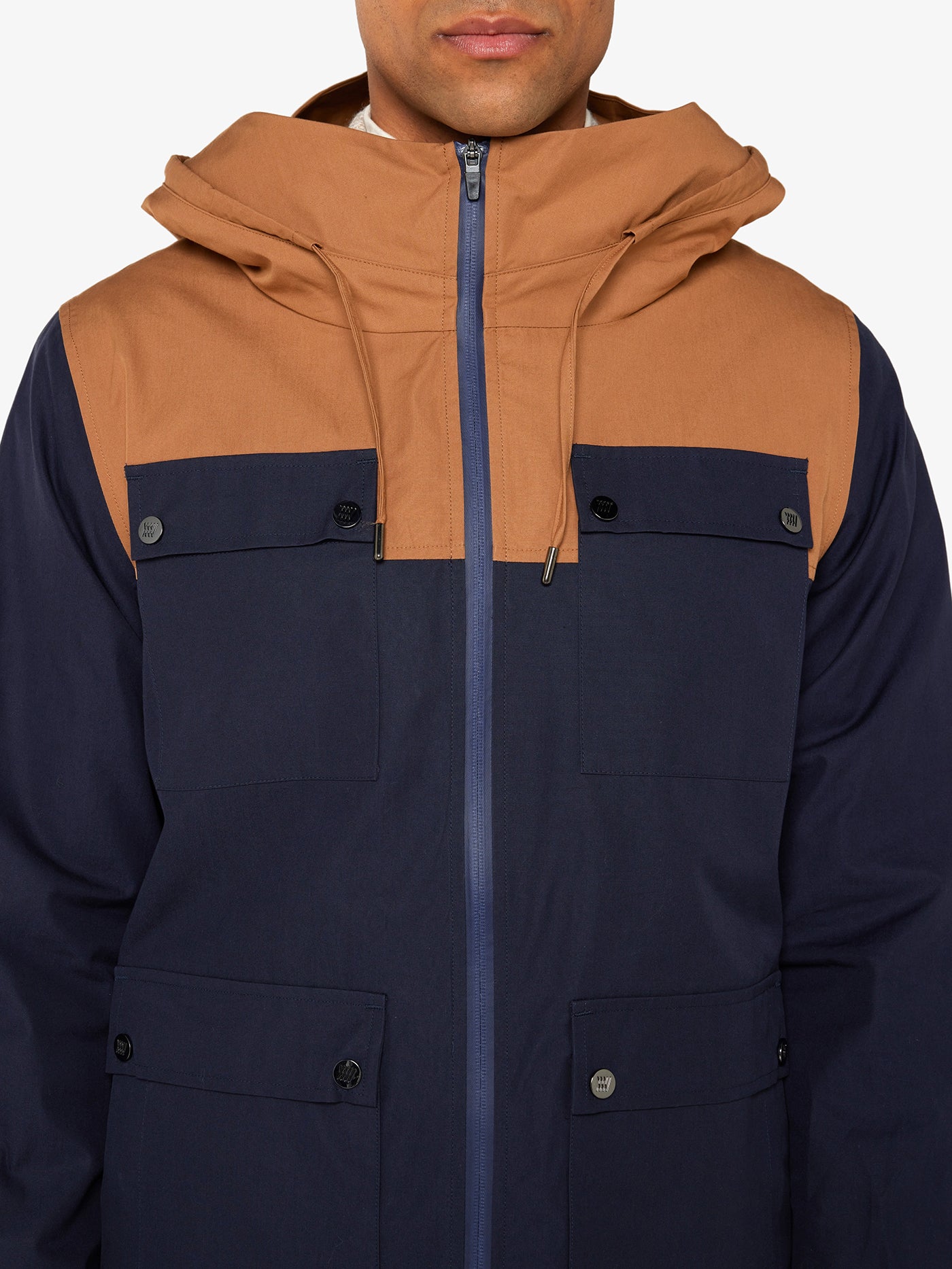 A person in a We Norwegians Lyngen Jacket Men Dark Blue, featuring Ventile® cotton with a brown hood/upper section and dark blue lower section. This weatherproof jacket has a front zipper, side pockets, and buttoned chest flap pockets for outdoor enthusiasts. The person's face is partially visible.