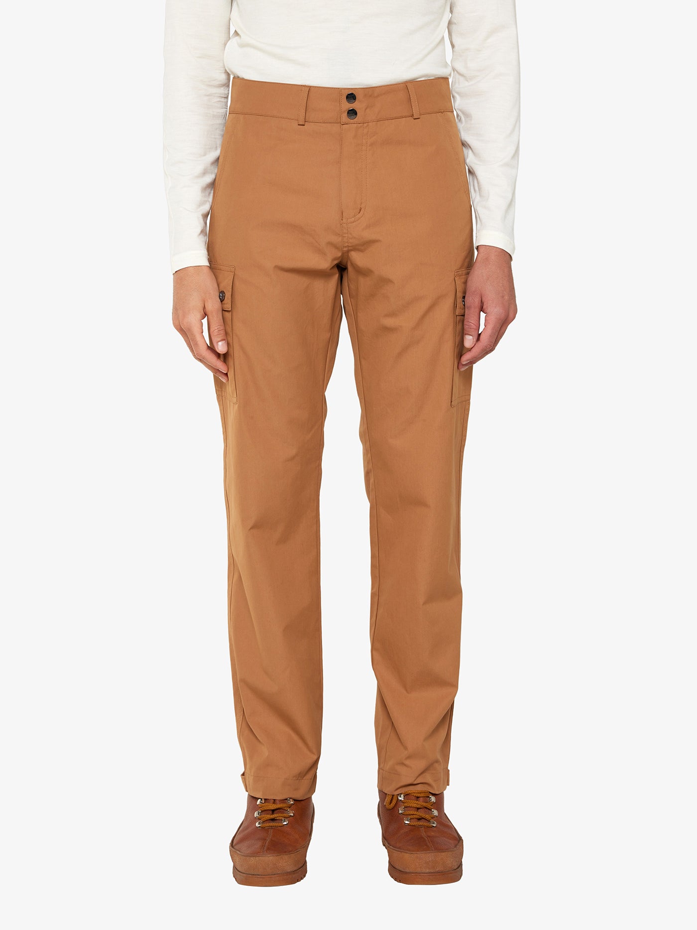 A person is wearing the Lyngen Pant Men Cinnamon from We Norwegians, which are brown cargo pants made from Ventile® cotton fabric, paired with a white long-sleeve shirt. The pants, ideal for outdoor enthusiasts with their unique weatherproof qualities and side pockets, are matched with brown lace-up boots. They stand against a plain white background.