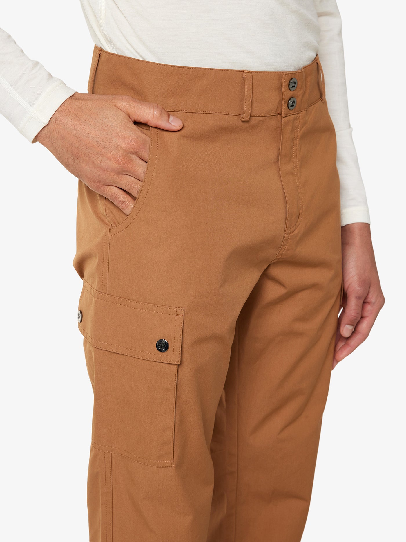 A person wearing a white, long-sleeved shirt and the Lyngen Pant Men Cinnamon by We Norwegians, made from Ventile® cotton fabric known for its unique weatherproof qualities, stands with their right hand in the pants' pocket. The background is plain white.
