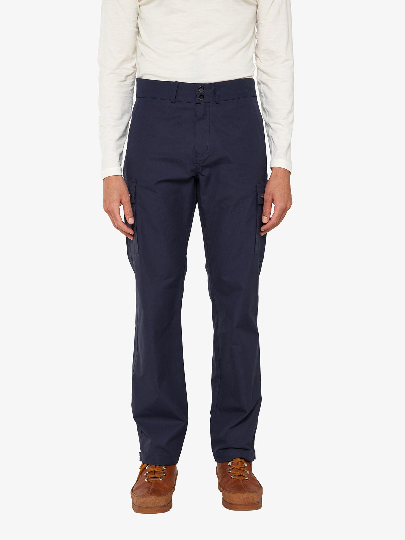 A person is wearing a white long-sleeve shirt made from Ventile® cotton fabric, We Norwegians' Lyngen Pant Men in Navy Blue, and brown leather shoes. The individual, clearly an outdoor enthusiast, is standing against a plain white background.