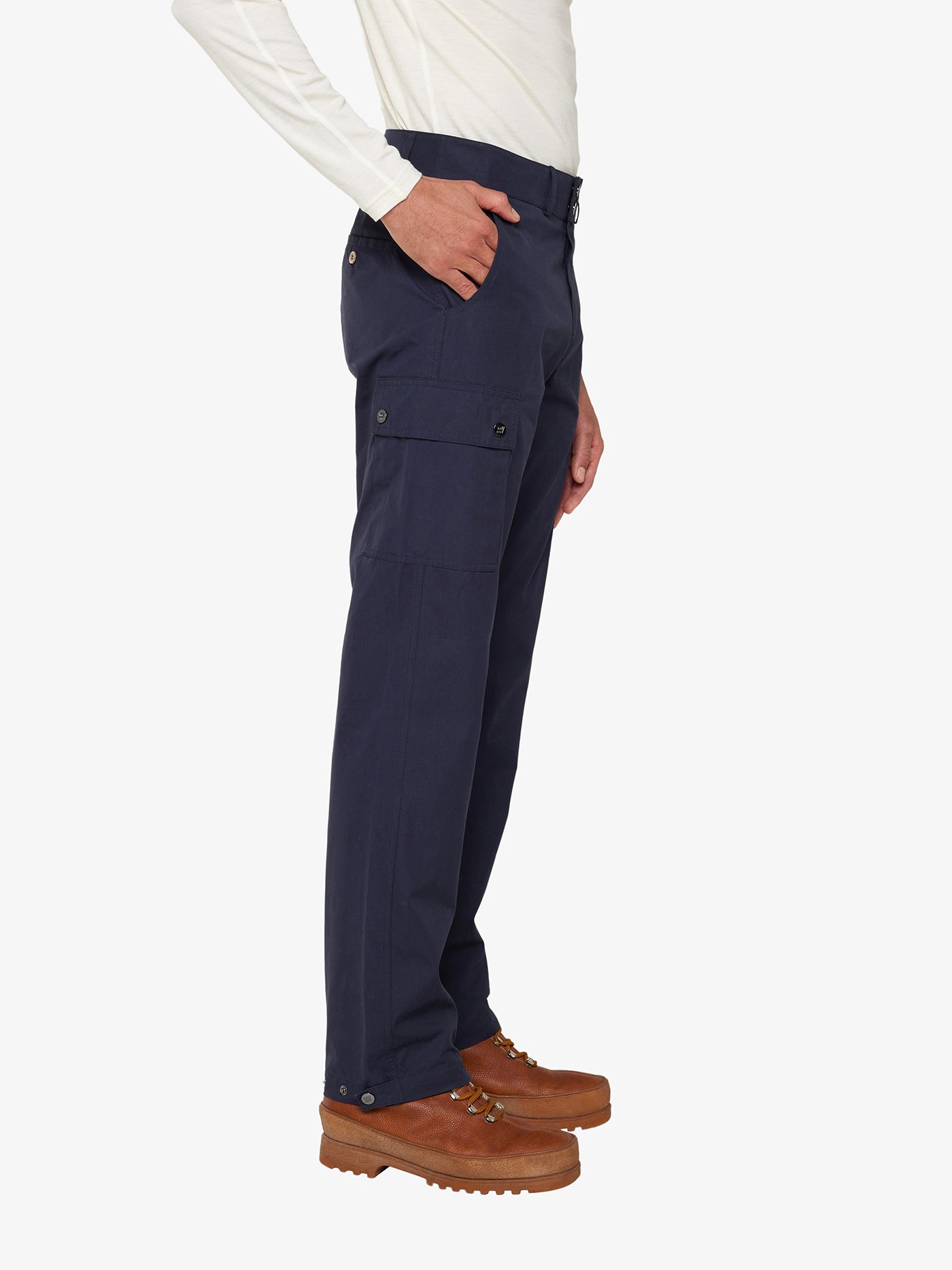 A person in a white long-sleeve shirt and brown boots is wearing the Lyngen Pant Men Navy Blue from We Norwegians, made from Ventile® cotton fabric. The weatherproof qualities of these pants, along with multiple pockets and side button details near the ankles, make them perfect for outdoor enthusiasts. The person is standing sideways with one hand in a pocket.