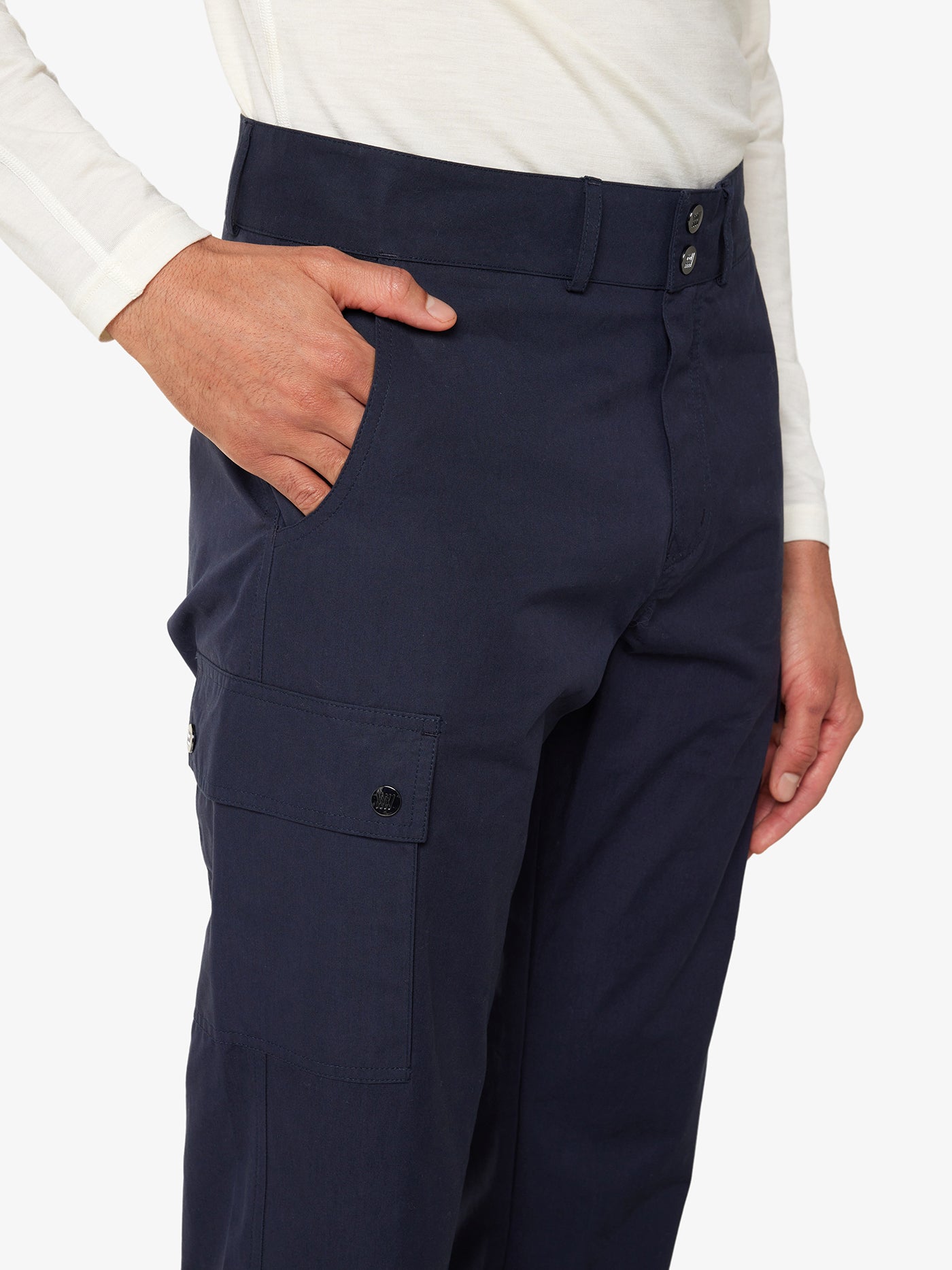 A person is shown wearing a white long-sleeved shirt and the We Norwegians Lyngen Pant Men Navy Blue, crafted from Ventile® cotton fabric. Their right hand is placed in their pants pocket. The pants, designed for outdoor enthusiasts, feature a buttoned side pocket and a double button and zipper closure at the waistband.