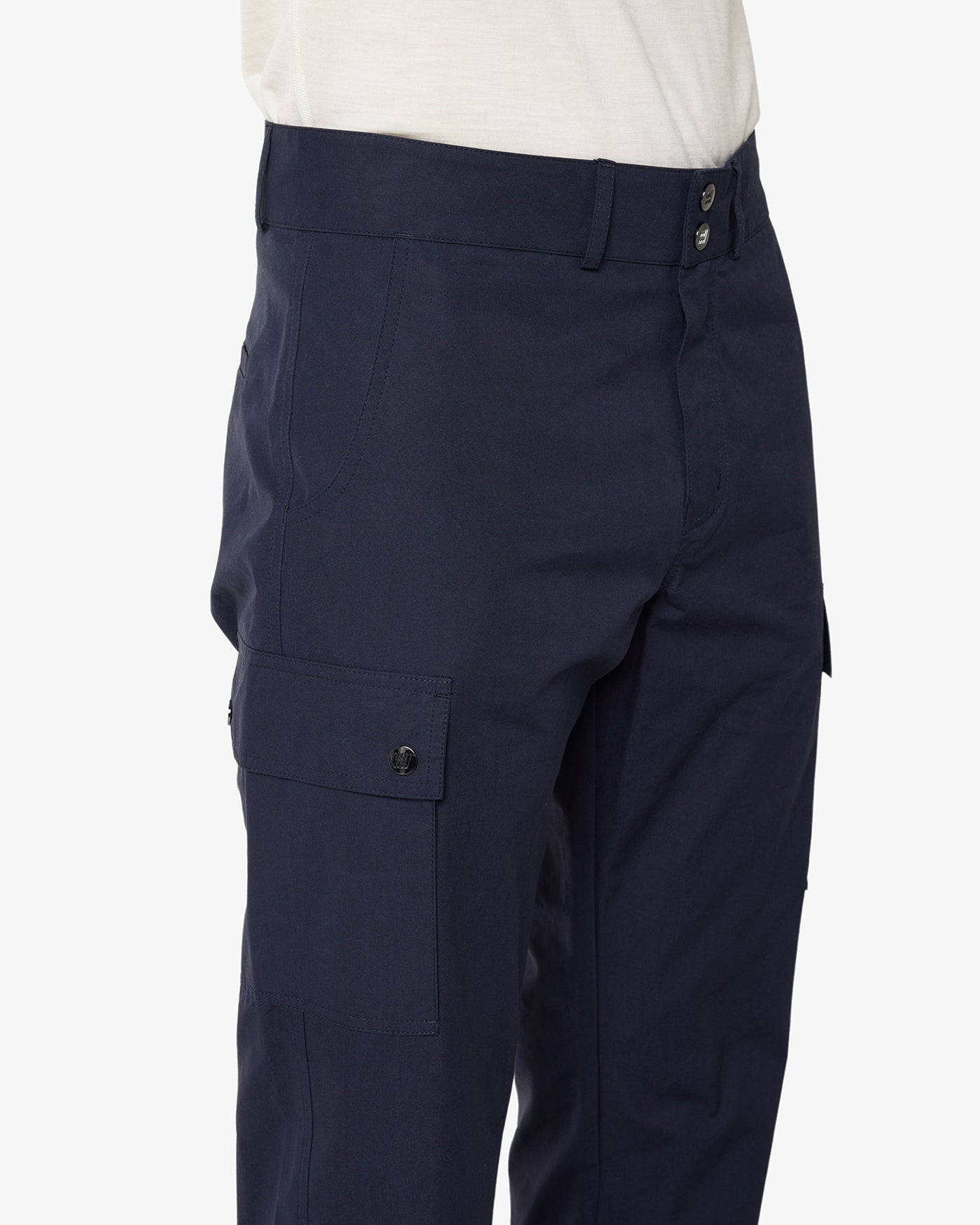 Close-up image of a person wearing the Lyngen Pant Men Navy Blue by We Norwegians, featuring multiple pockets. These dark blue cargo pants, made from Ventile® cotton fabric, have a button closure and are paired with a white shirt. Their weatherproof qualities ensure a snug fit around the waist, with visible seams and sturdy fabric—ideal for outdoor enthusiasts.