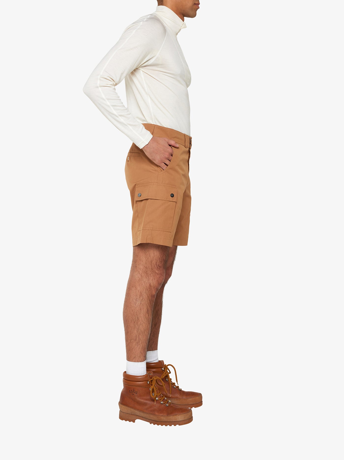 A person is shown from the side, wearing a white long-sleeve top tucked into We Norwegians' Lyngen Shorts Men Cinnamon, made from durable Ventile® fabric with pockets, paired with tan lace-up boots. The ensemble is perfect for outdoor enthusiasts and stands out against the plain white background.