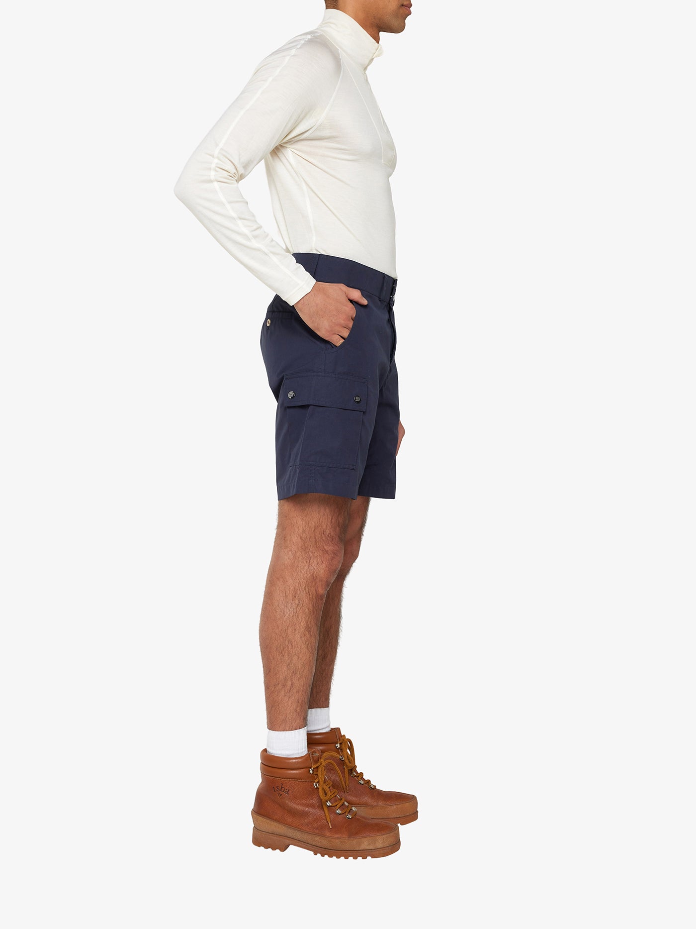 A person is standing in profile, wearing a long-sleeved white shirt made from weatherproof Ventile® cotton fabric, paired with Lyngen Shorts Men Navy Blue by We Norwegians and brown lace-up boots with white socks. The person has their hands in the pockets of the shorts against a plain white background.