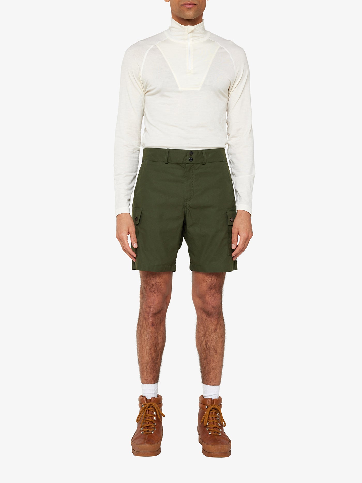 A person is wearing a long-sleeved, cream-colored top made from Ventile® cotton fabric with a half-zip neckline and stands with their hands by their sides. They are also wearing Lyngen Shorts Men in Olive Green by We Norwegians, white socks, and brown lace-up hiking boots. The background is plain white.