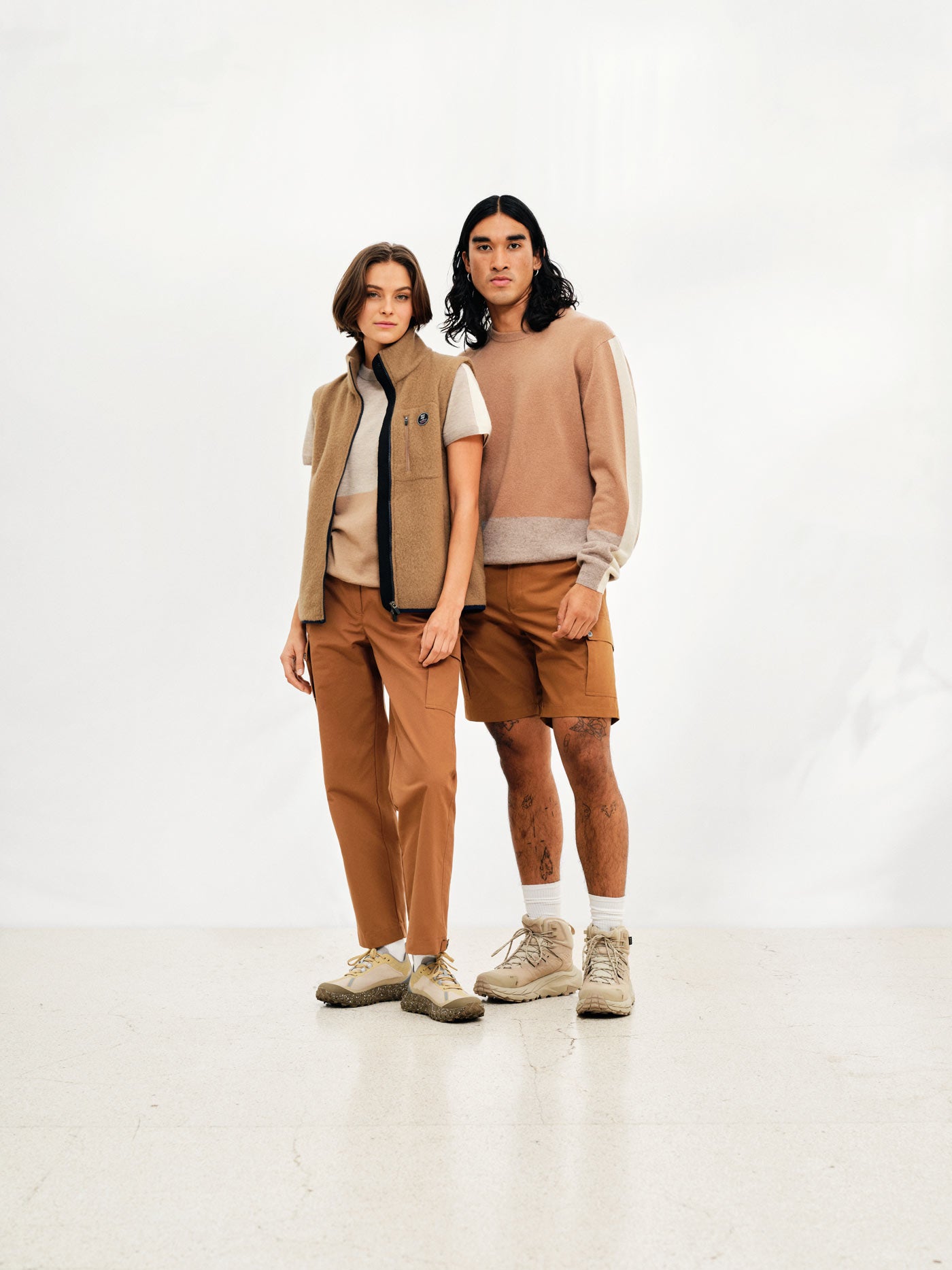 Two individuals pose against a white background. The person on the left wears a beige vest, light brown pants, and white sneakers. The person on the right dons a beige sweater, Lyngen Shorts Men in Cinnamon by We Norwegians, and white sneakers. Both have serious expressions and neutral postures.