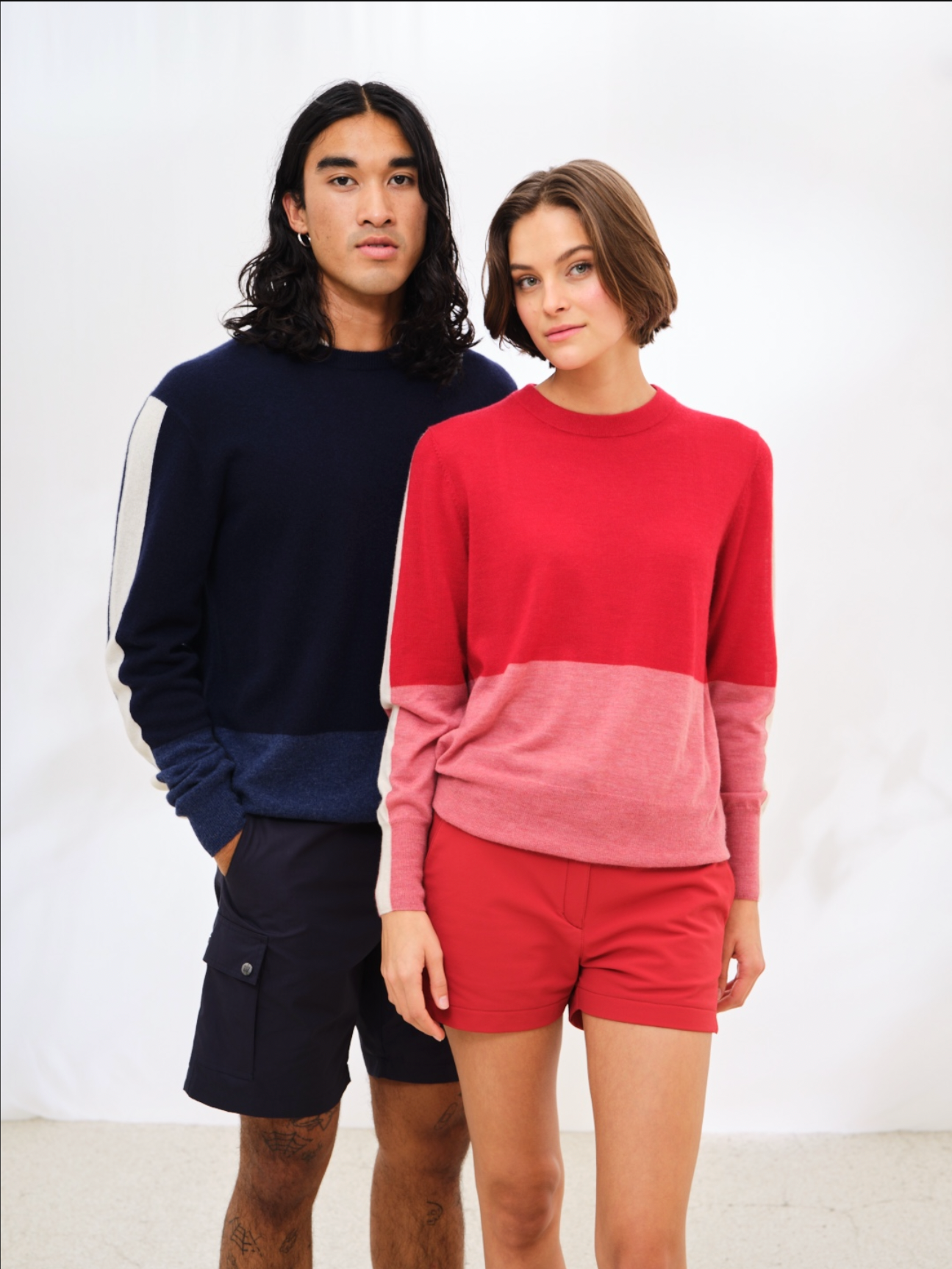 Two people stand side by side against a white background. The person on the left, with long dark hair, is wearing a navy blue and white sweater paired with We Norwegians' Lyngen Shorts Men Navy Blue made from Ventile® cotton fabric. The person on the right has shoulder-length light brown hair, a red and pink sweater, and red shorts.