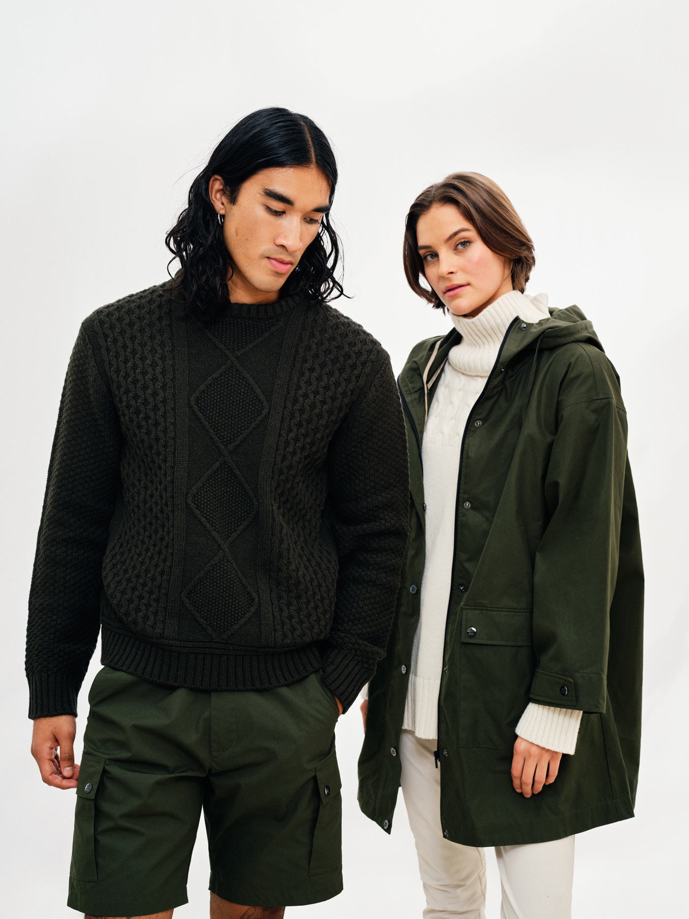 A man and a woman stand side by side against a white background. The man has long black hair, wears a dark green textured sweater and the Lyngen Shorts Men Olive Green by We Norwegians. The woman, reminiscent of RAF pilots with her weatherproof qualities, has short brown hair and dons a white turtleneck under a dark green hooded coat crafted from Ventile® cotton fabric.