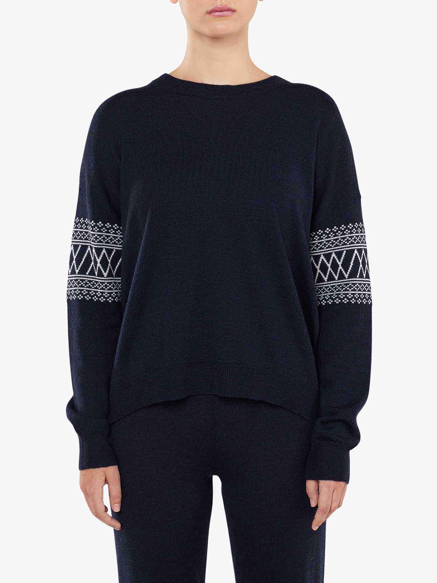 A person wearing We Norwegians' Signature Sweater Women, characterized by its relaxed fit and intricate white Setesdal patterns on the sleeves in dark blue Merino wool, paired with matching dark blue pants. The background is plain white.