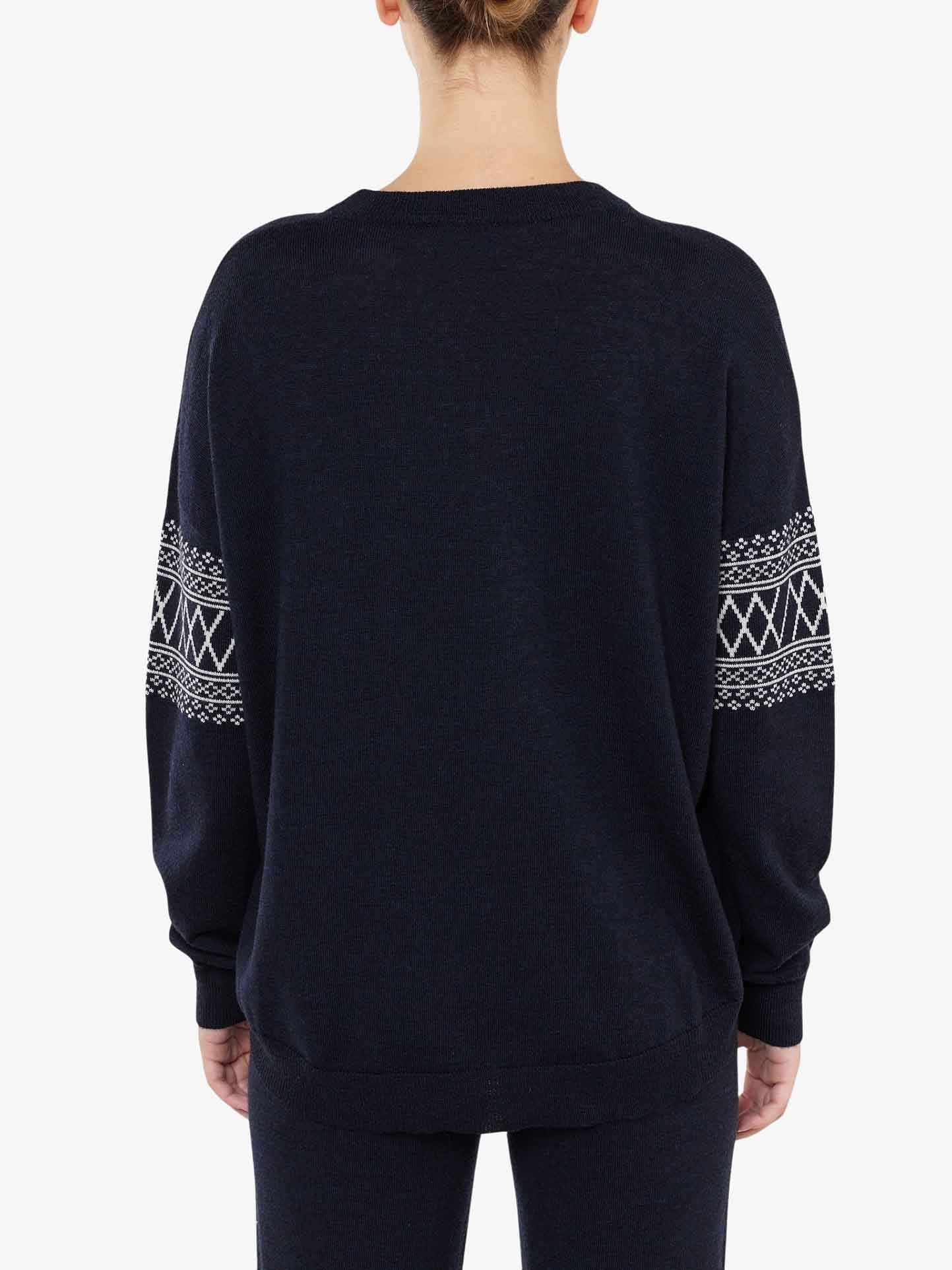 A person with hair tied back is seen from behind, wearing an over-sized black Signature Sweater Women by We Norwegians, featuring intricate white Setesdal patterns on the sleeves. The Merino wool sweater has a loose, comfortable fit and the person is also wearing matching black pants. The background is plain white.