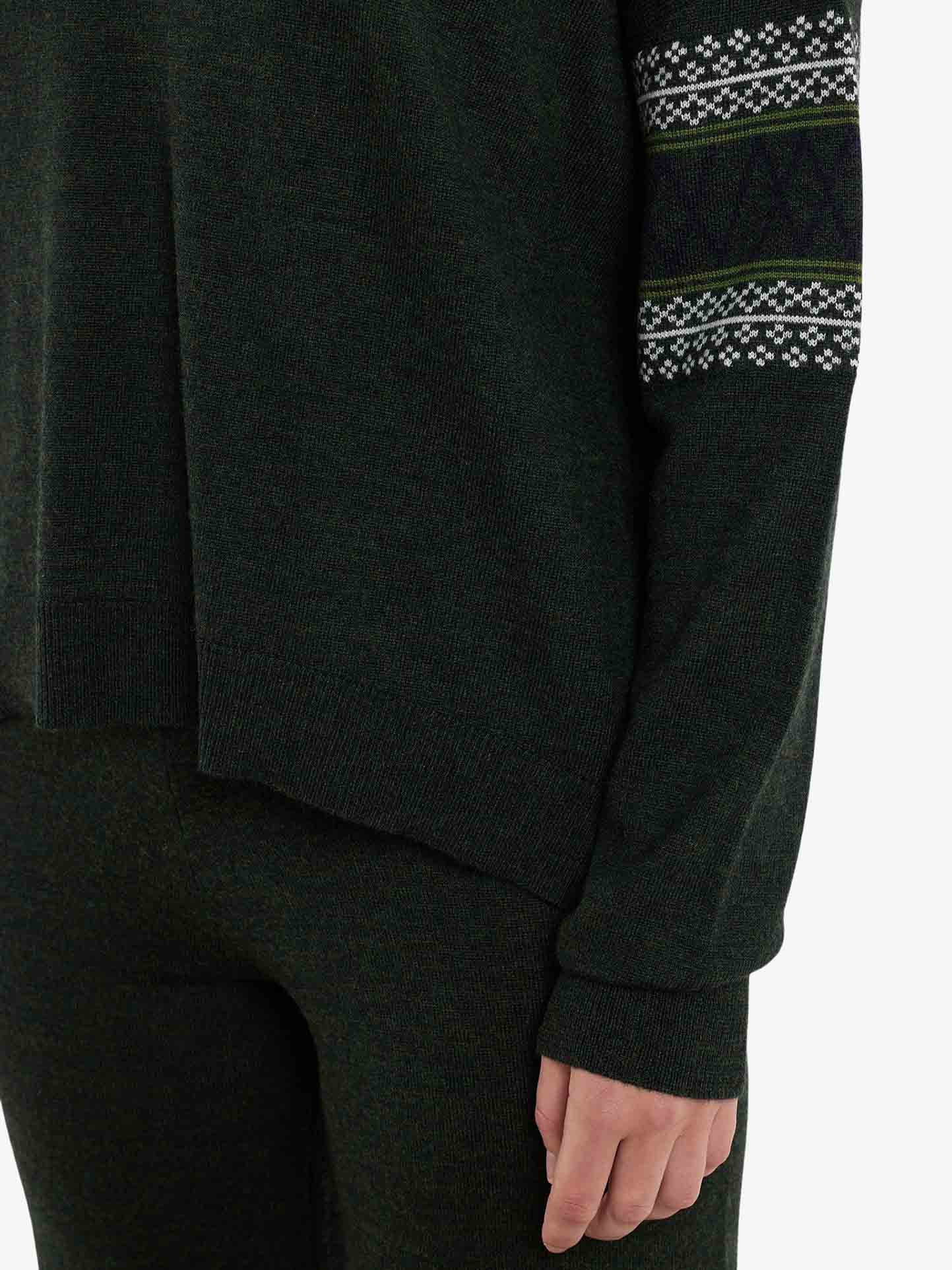 A person is wearing the Signature Sweater Women from We Norwegians in dark green, paired with matching pants. This over-sized sweater, crafted from Merino wool, showcases a patterned design with white geometric shapes reminiscent of the Setesdal pattern and green stripes on the sleeves. The background is plain white. The lower part of the face and entire upper body are not visible.
