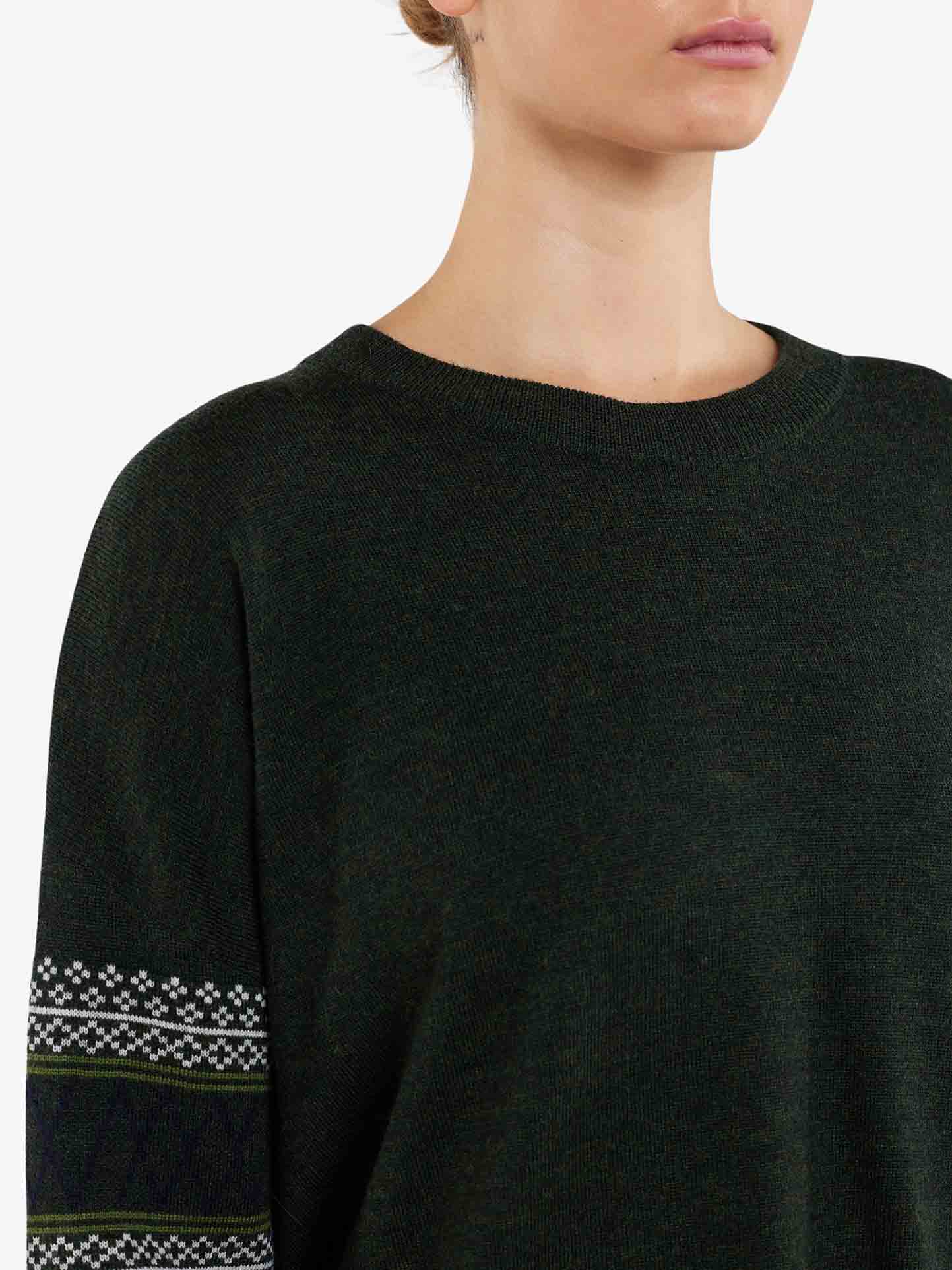 Close-up of a person wearing an over-sized Signature Sweater Women by We Norwegians in dark green, featuring patterned sleeves. The intricate Setesdal pattern includes horizontal stripes of white interspersed with smaller green and black designs. The person’s head is partially out of frame, and their hair is tied back.