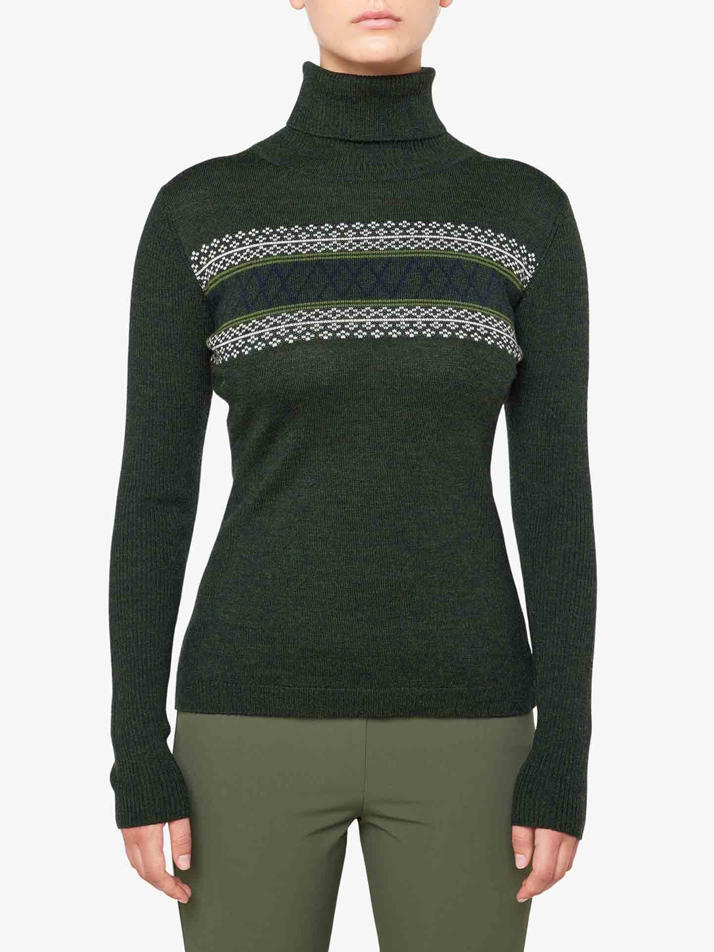 A person is wearing the We Norwegians Signature Rollneck Women sweater in olive green, crafted from fine Merino wool with a patterned knit design across the chest. The sweater boasts long sleeves, and the individual pairs it with matching olive green pants. The plain white background enhances the cozy, retro ski outfit vibes.