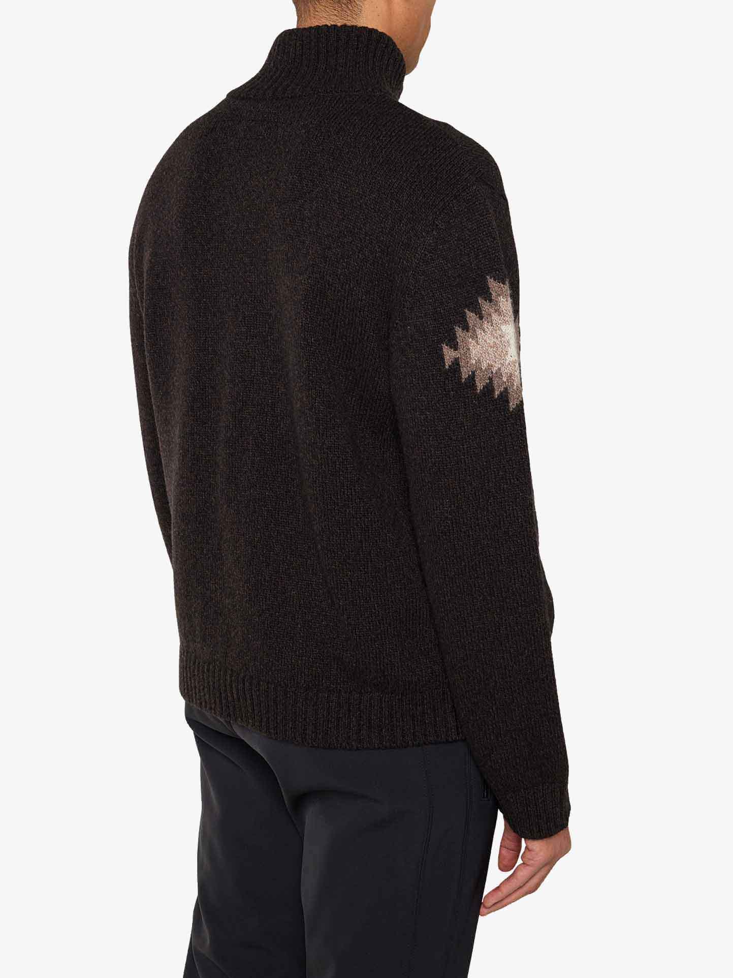 Rear view of a person wearing the Hovden Sweater Men by We Norwegians, featuring a black turtleneck design with a white and brown geometric pattern on one sleeve that evokes Norwegian Sámi cultural motifs, paired with dark trousers—perfect for Fall weather.