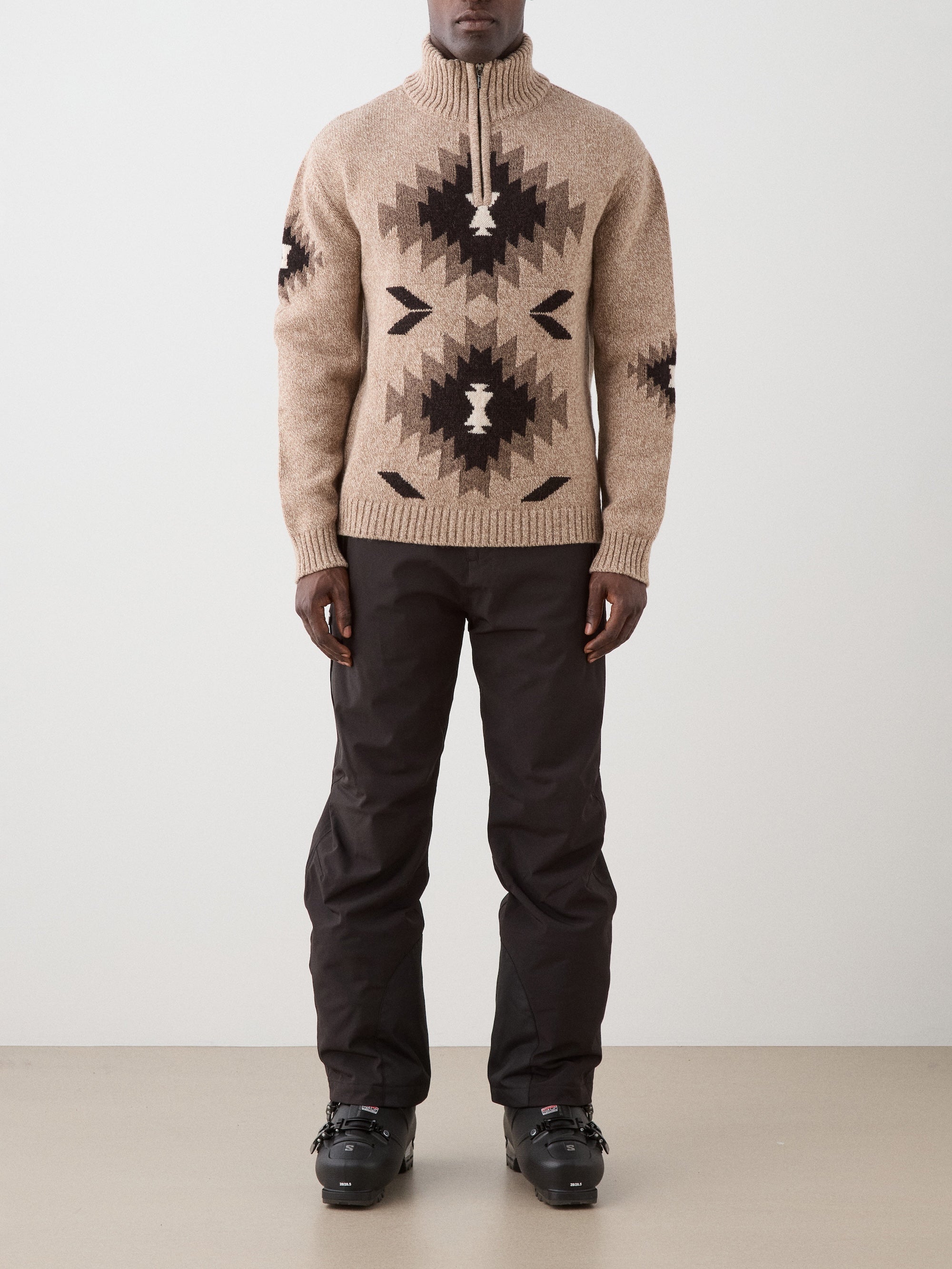 A man wearing the We Norwegians Hovden Sweater for men—beige with black geometric patterns—paired with black pants and winter boots, stands facing forward against a plain light-colored wall, hands at his sides.